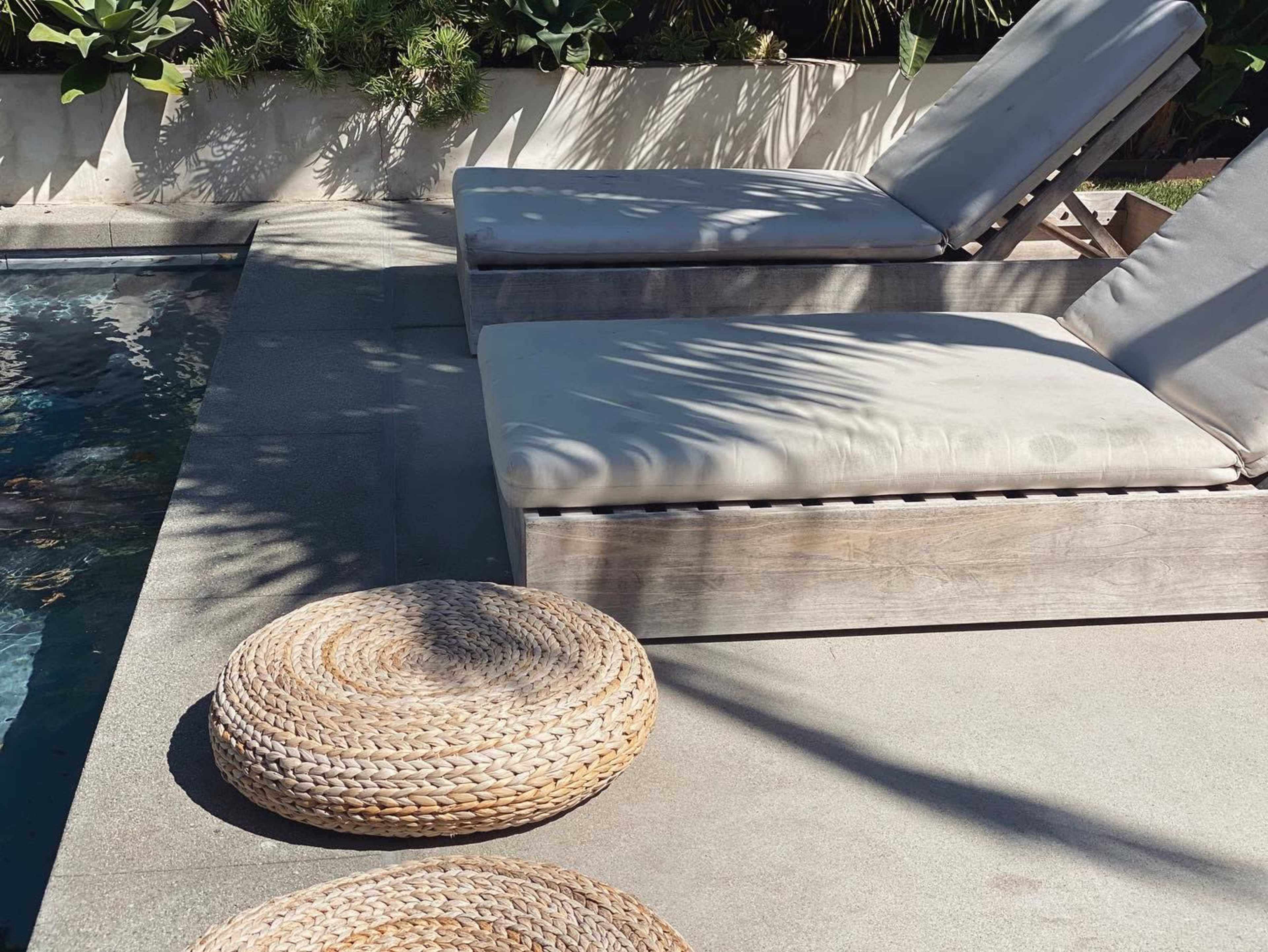 Two gray lounge chairs are positioned beside a swimming pool, accompanied by a round woven mat on the concrete surface.