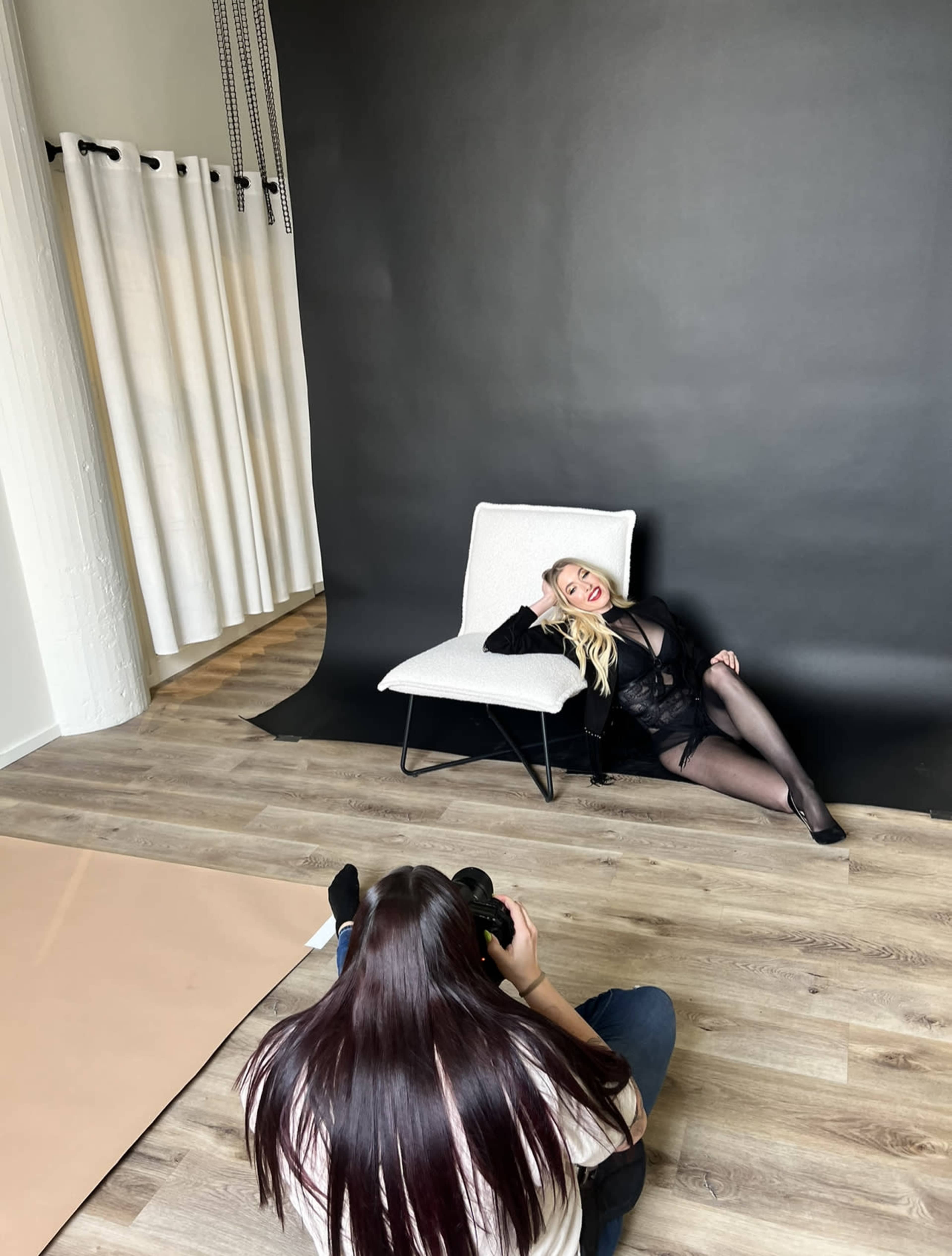 A model poses on a chair against a black backdrop while a photographer captures the scene from the floor.