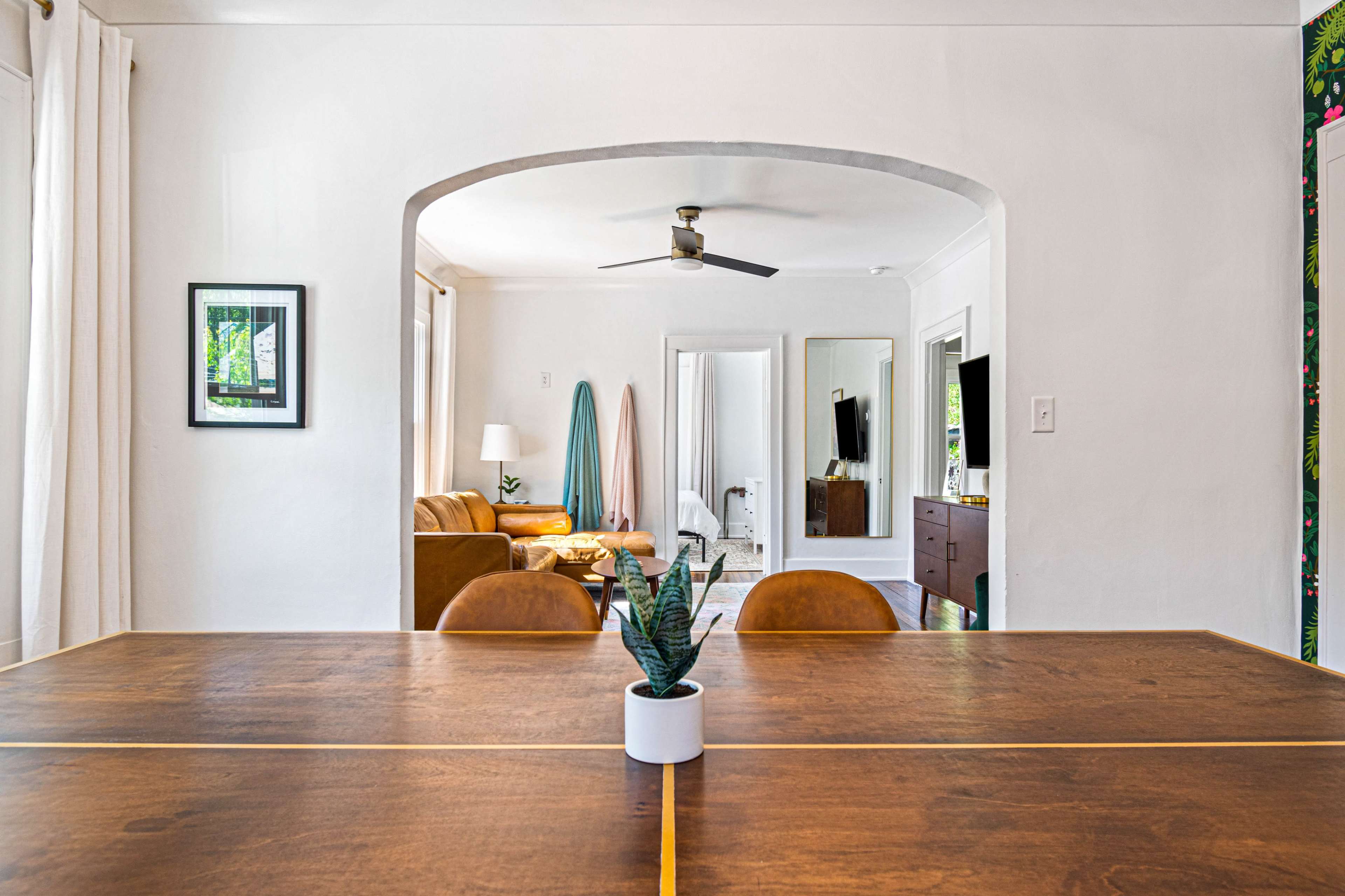 A spacious dining area with a wooden table and a small potted plant, leading to a living room with a brown sofa and mirrors.