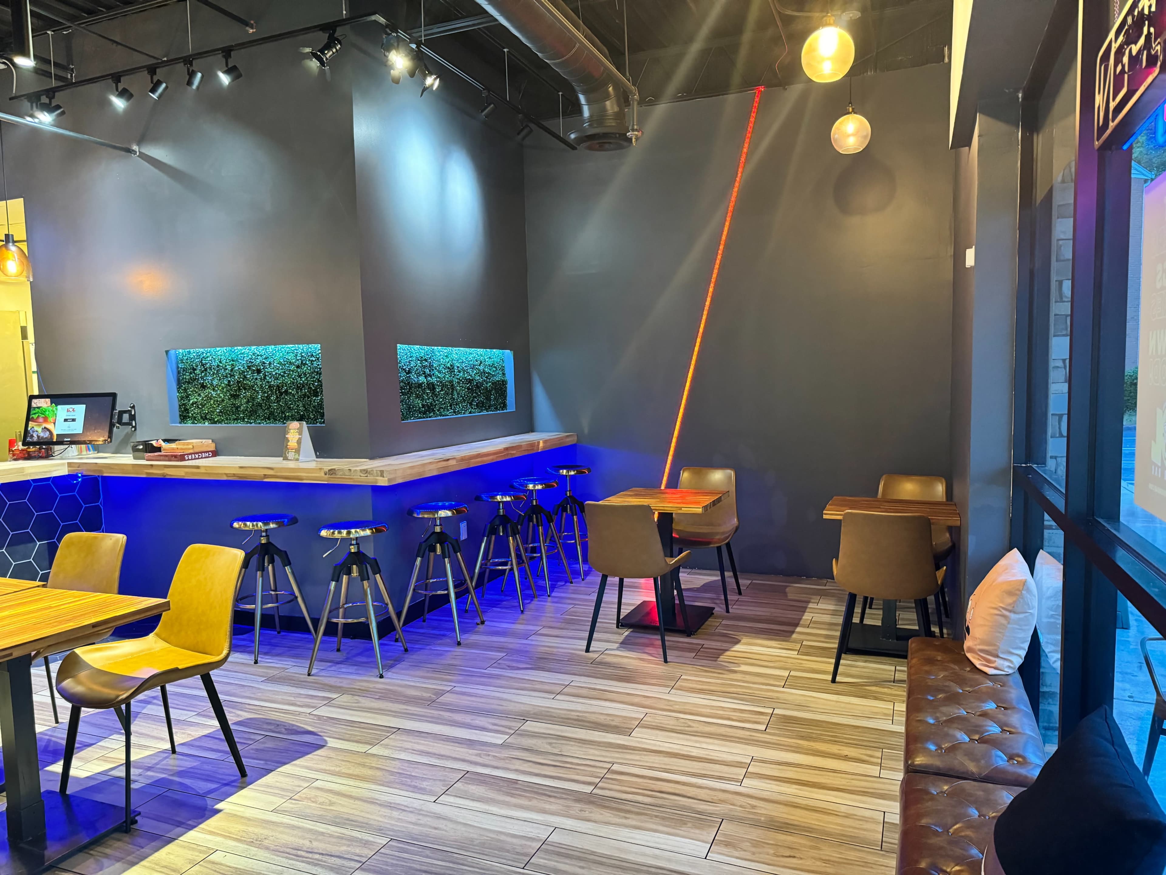 A modern cafe interior featuring wooden tables, metallic stools, and a softly lit environment with a green plant wall and blue ambient lighting.