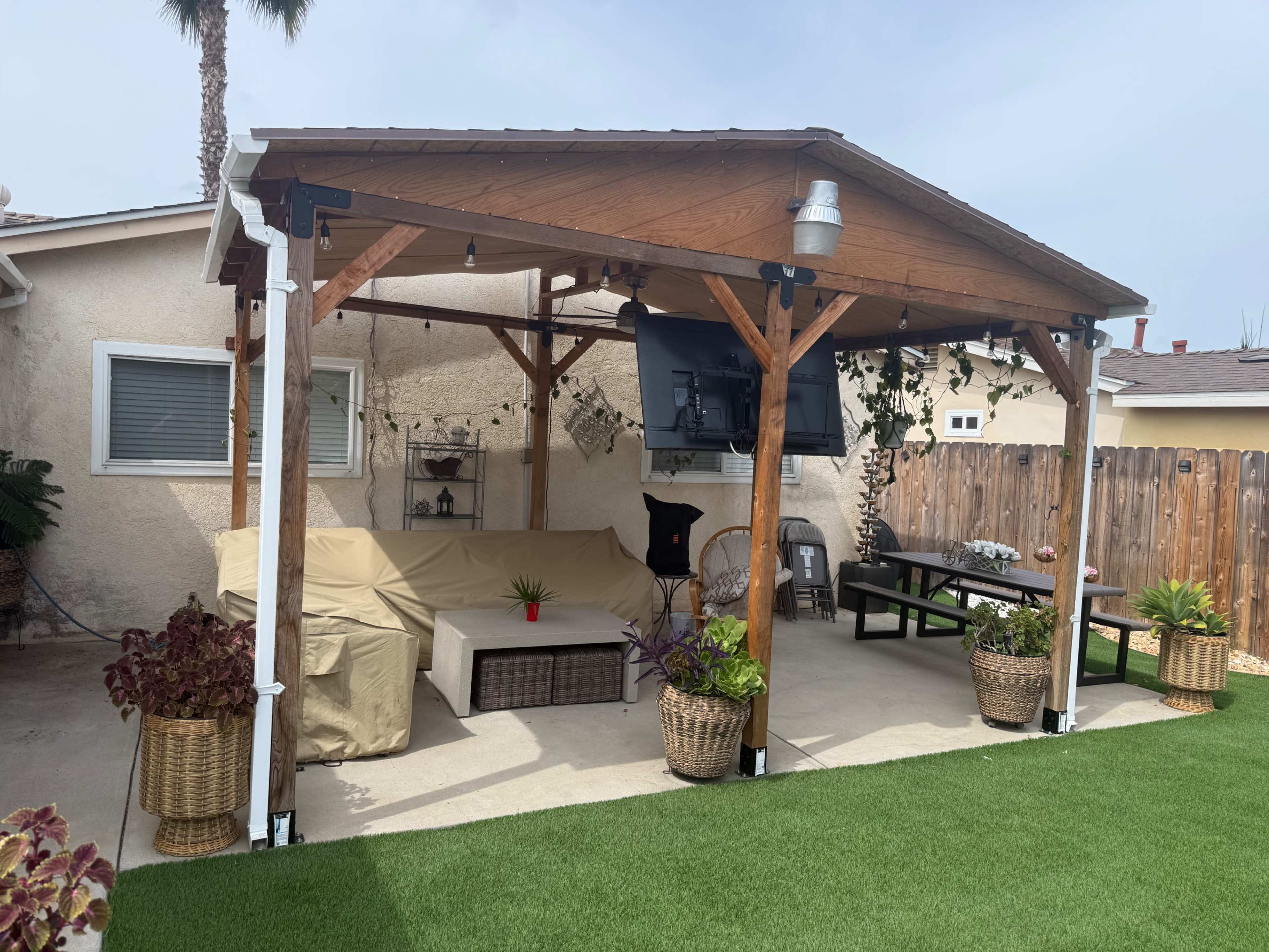The image shows a covered outdoor patio with wooden beams, a sofa covered with a protective tarp, a television mounted on the wall, and a dining table on a green lawn.