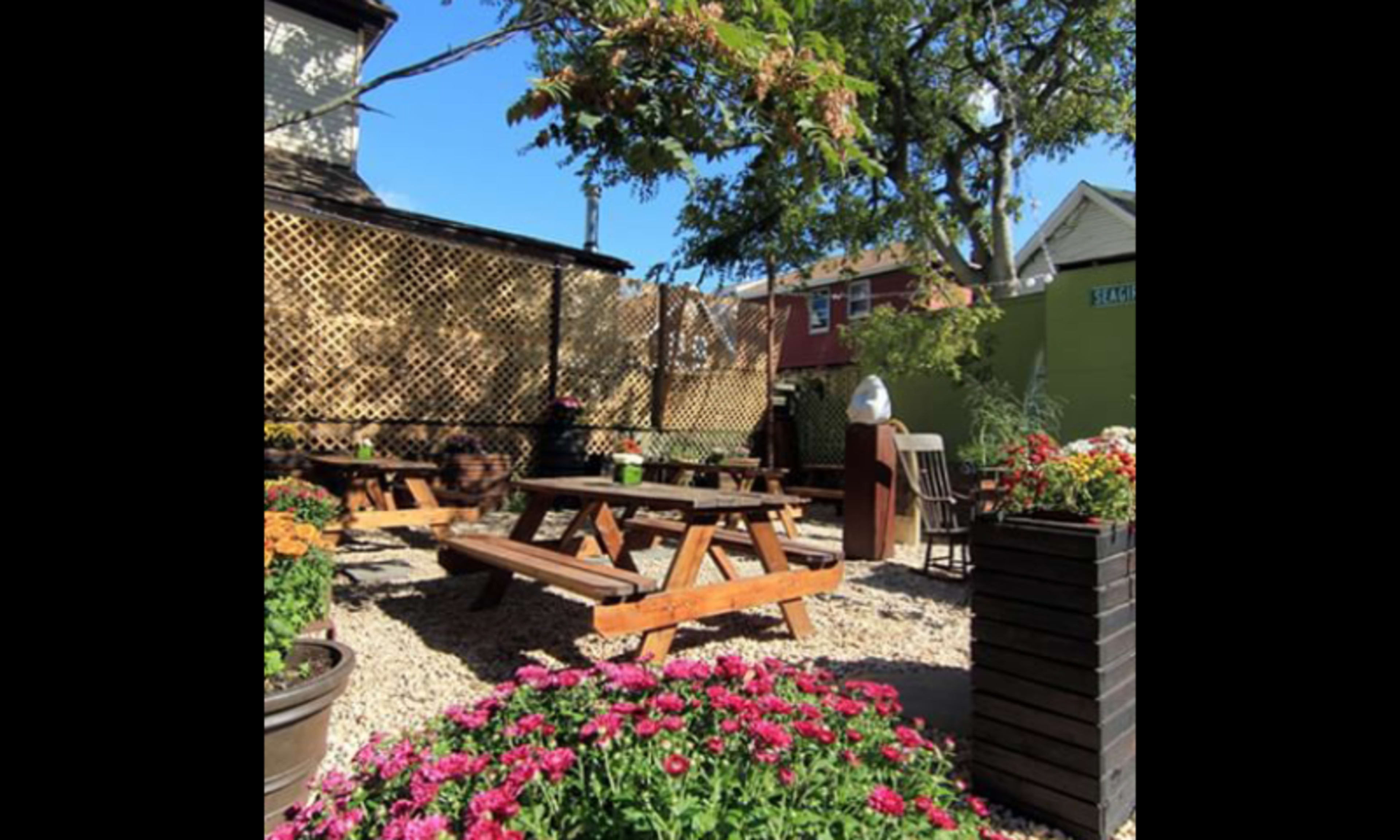 An outdoor patio area with wooden picnic tables surrounded by flowers and a grill, enclosed by lattice fencing and buildings.