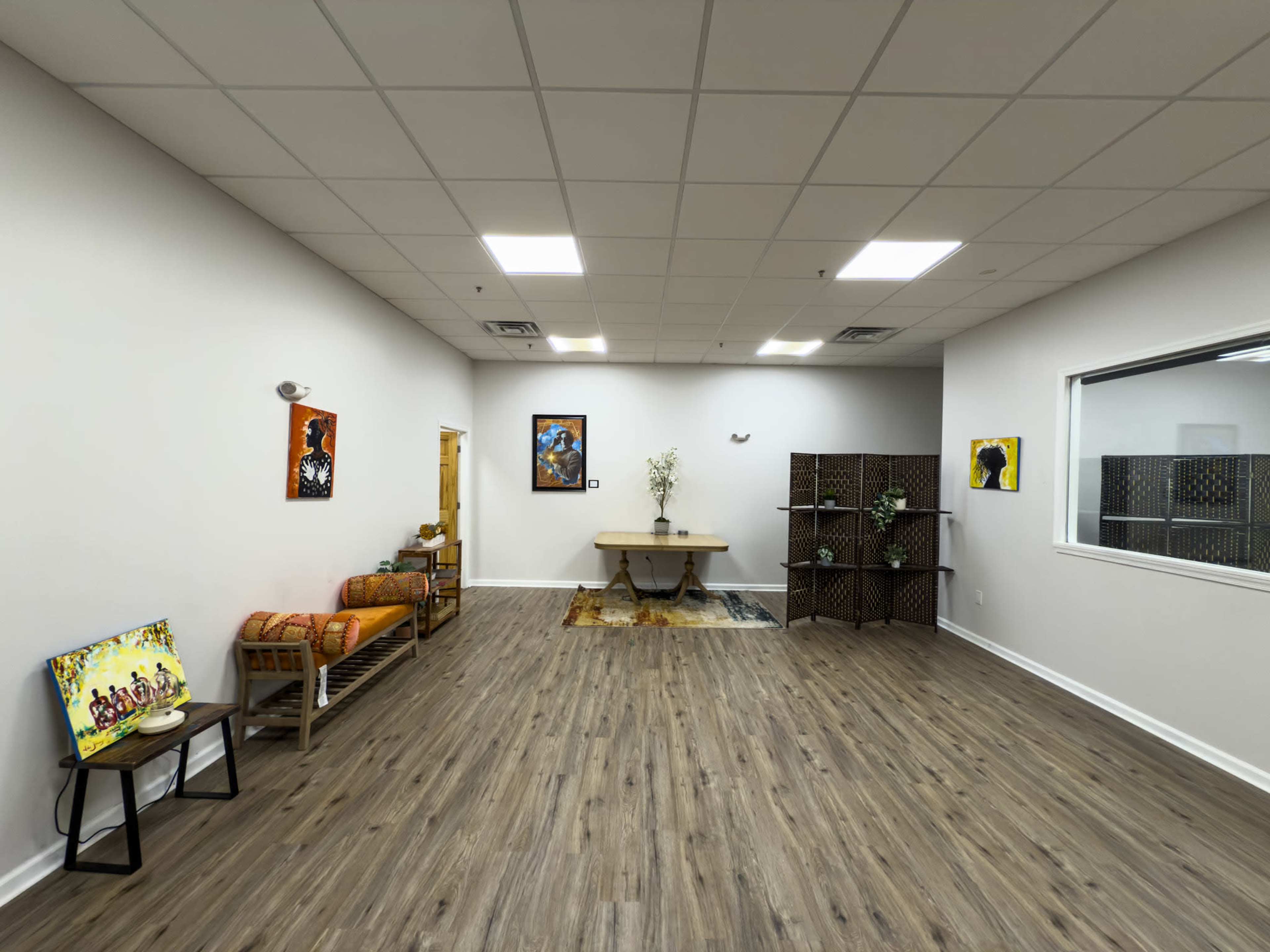 The image shows a spacious, well-lit room featuring a wooden table at the center, surrounded by minimal furniture and artwork on the walls.