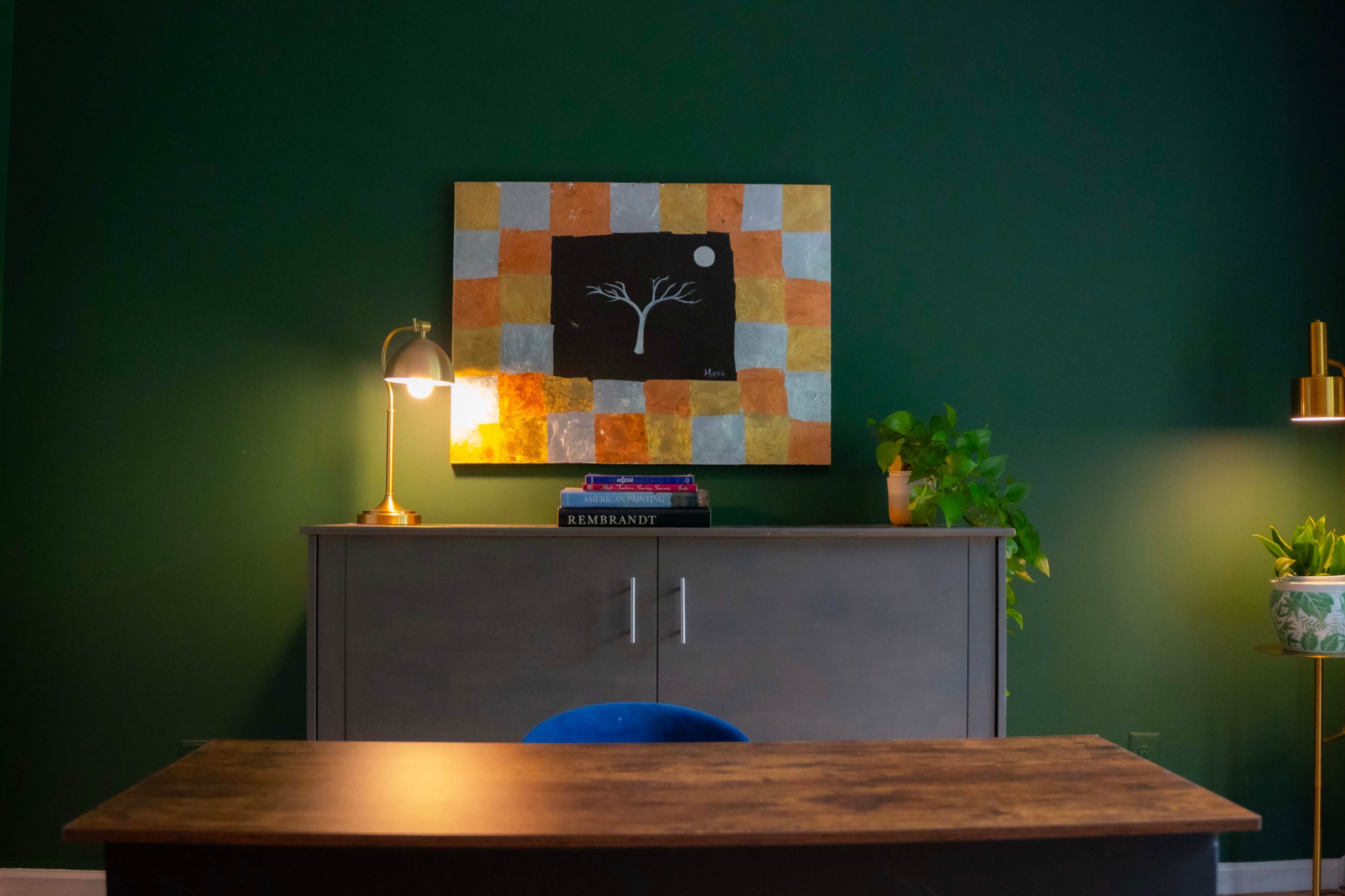 A dark green wall features an abstract painting above a wooden cabinet, with a lamp and plants positioned nearby.