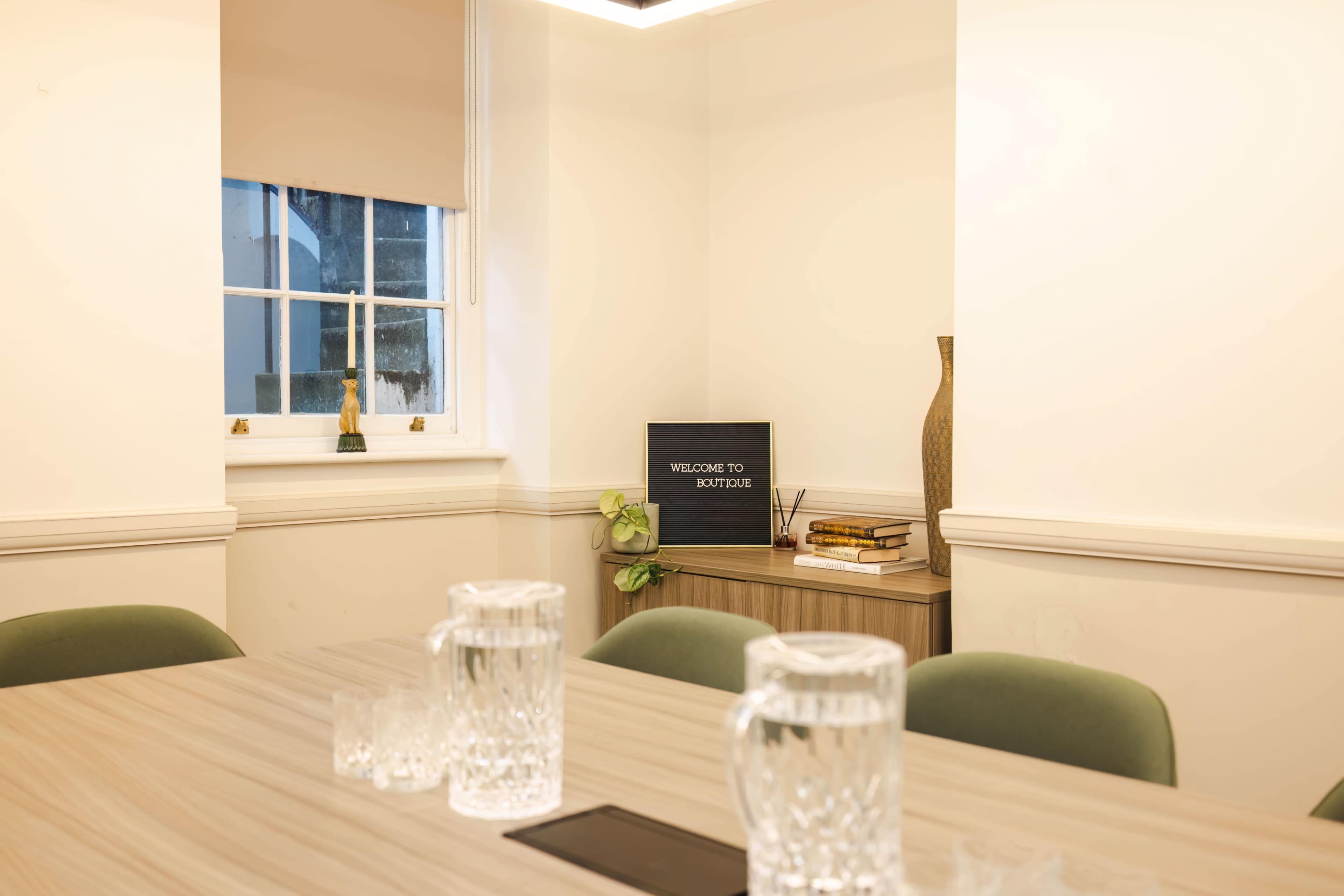 A modern meeting room features a wooden table with glass pitchers and a welcome sign on a side cabinet.