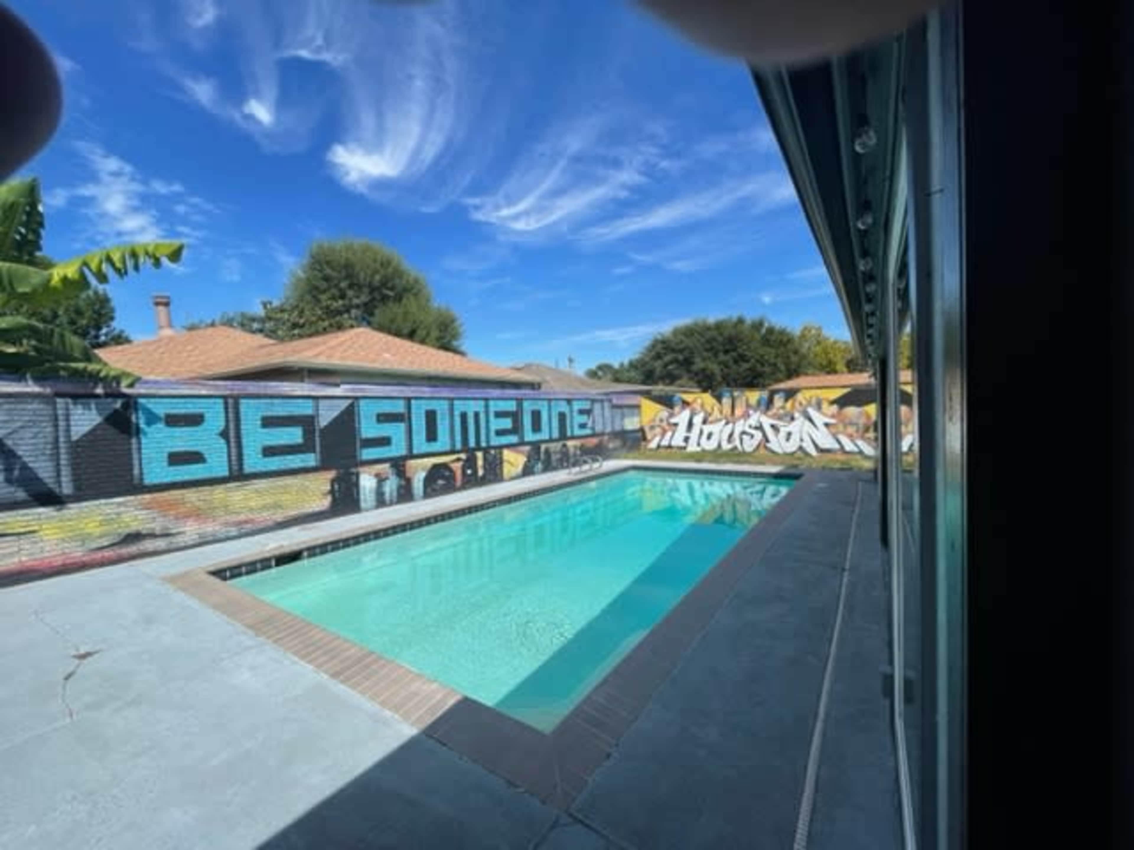 A swimming pool surrounded by a modern home, featuring a vibrant mural on the wall that reads "BE SOMEONE" against a blue sky.