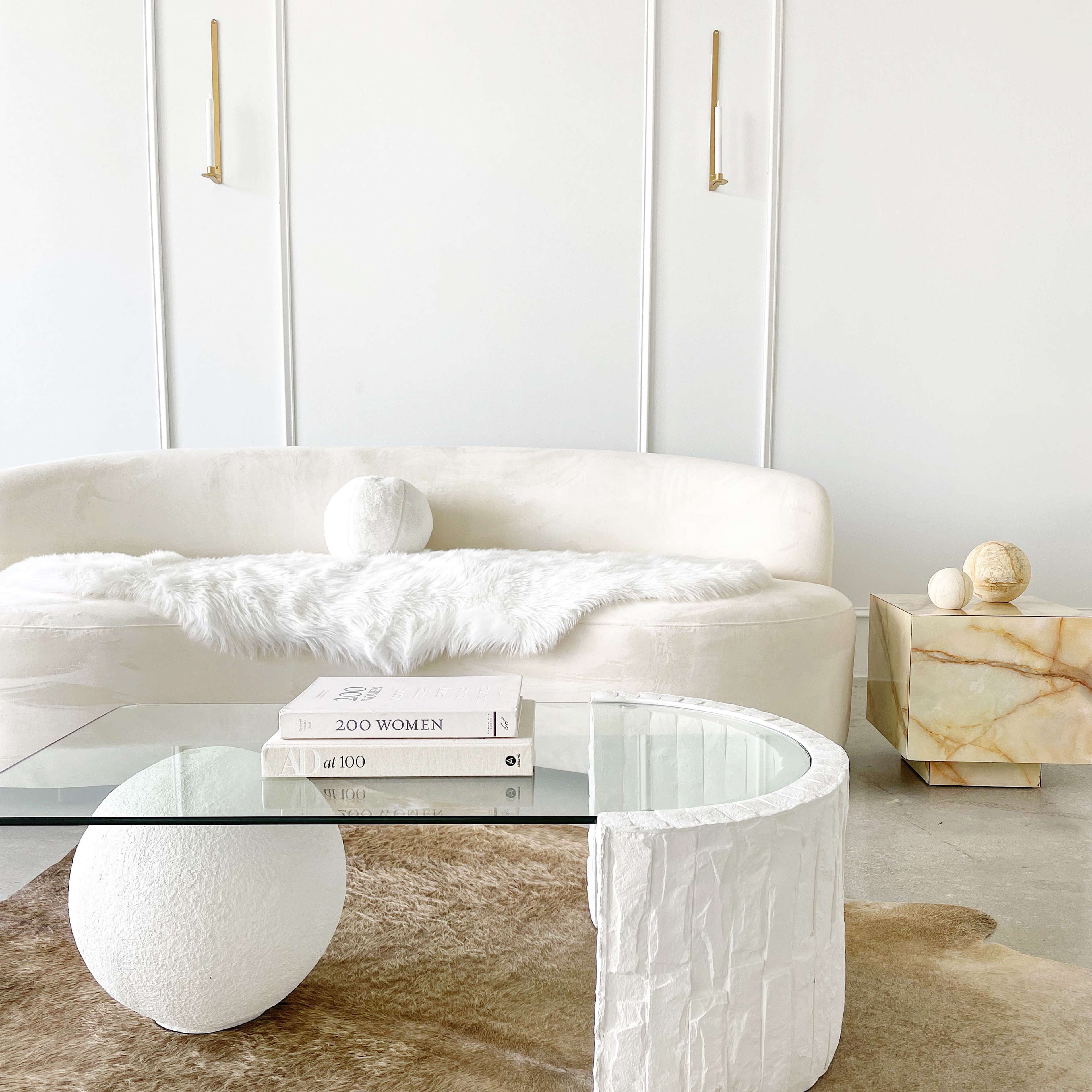 A minimalist living room features a round white couch, a glass coffee table with stacked books, and a decorative stone sphere on a textured rug.