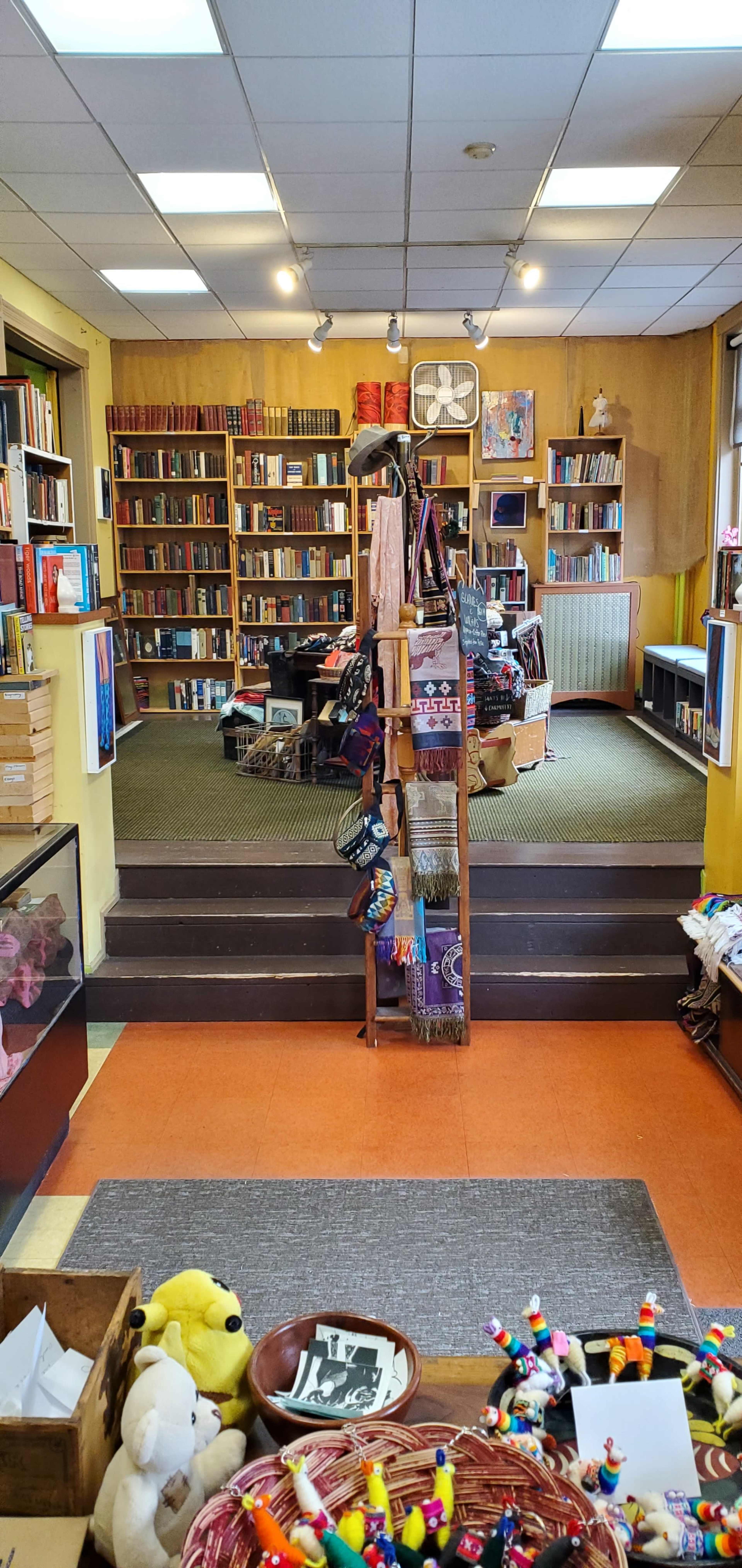 A cozy bookstore interior with wooden bookshelves, a staircase leading to a reading area, and various items for sale in the foreground.