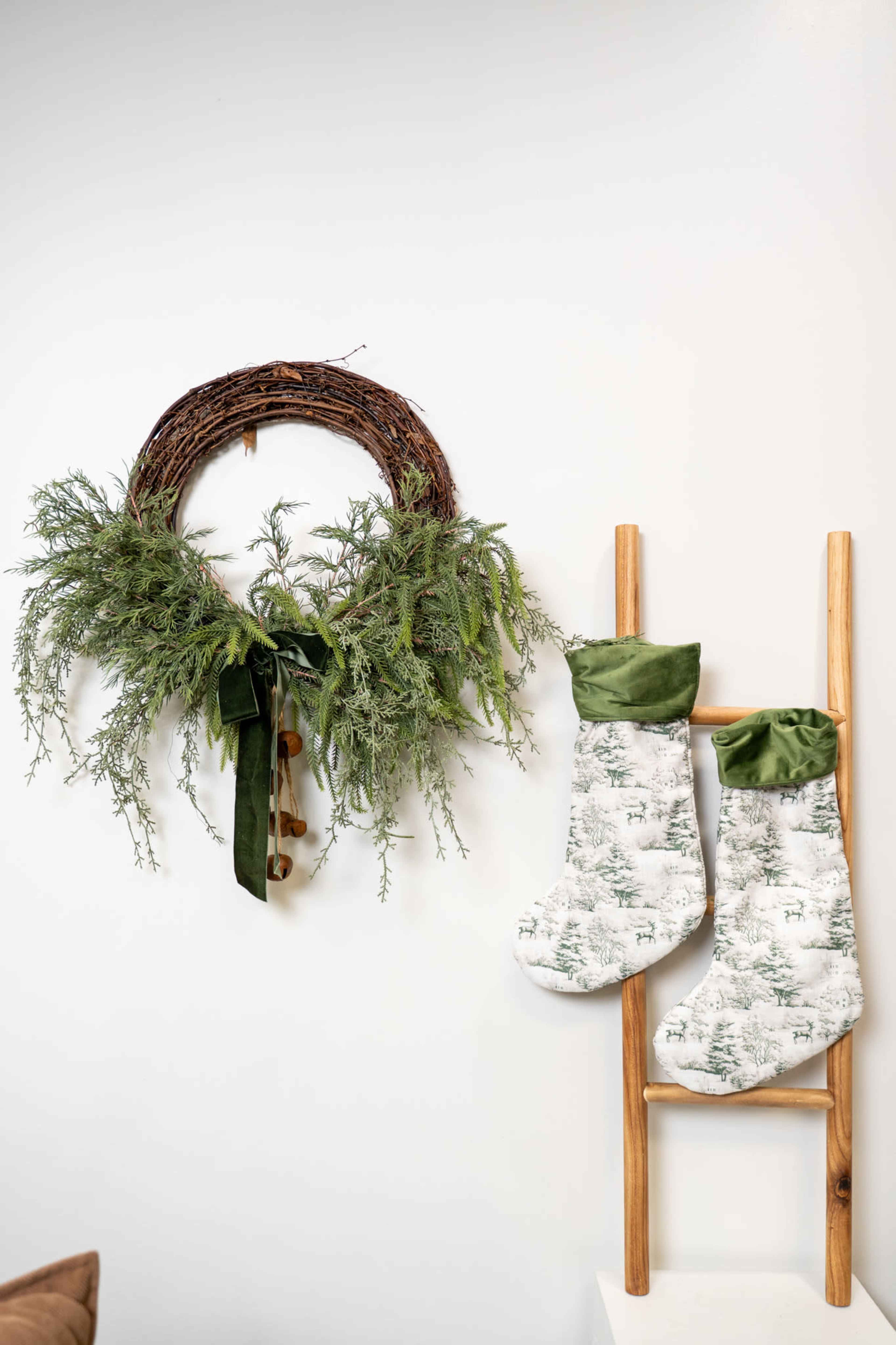 A circular grapevine wreath adorned with greenery and a ribbon is displayed on a wall next to two hanging Christmas stockings on a wooden ladder.