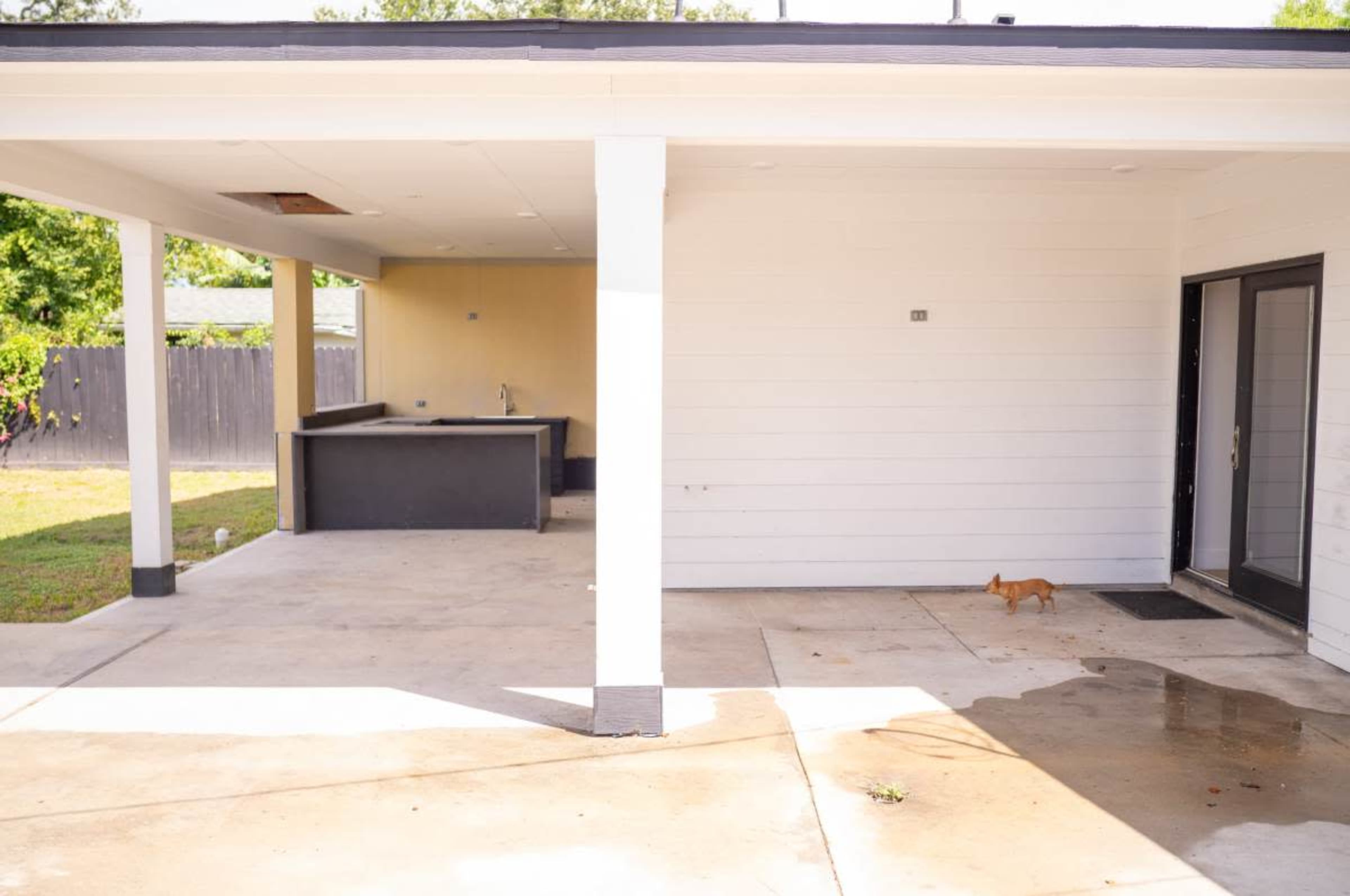 The image shows a covered patio area with a small outdoor kitchen space on one side and a single story white house wall on the other, with a small dog walking on the concrete.