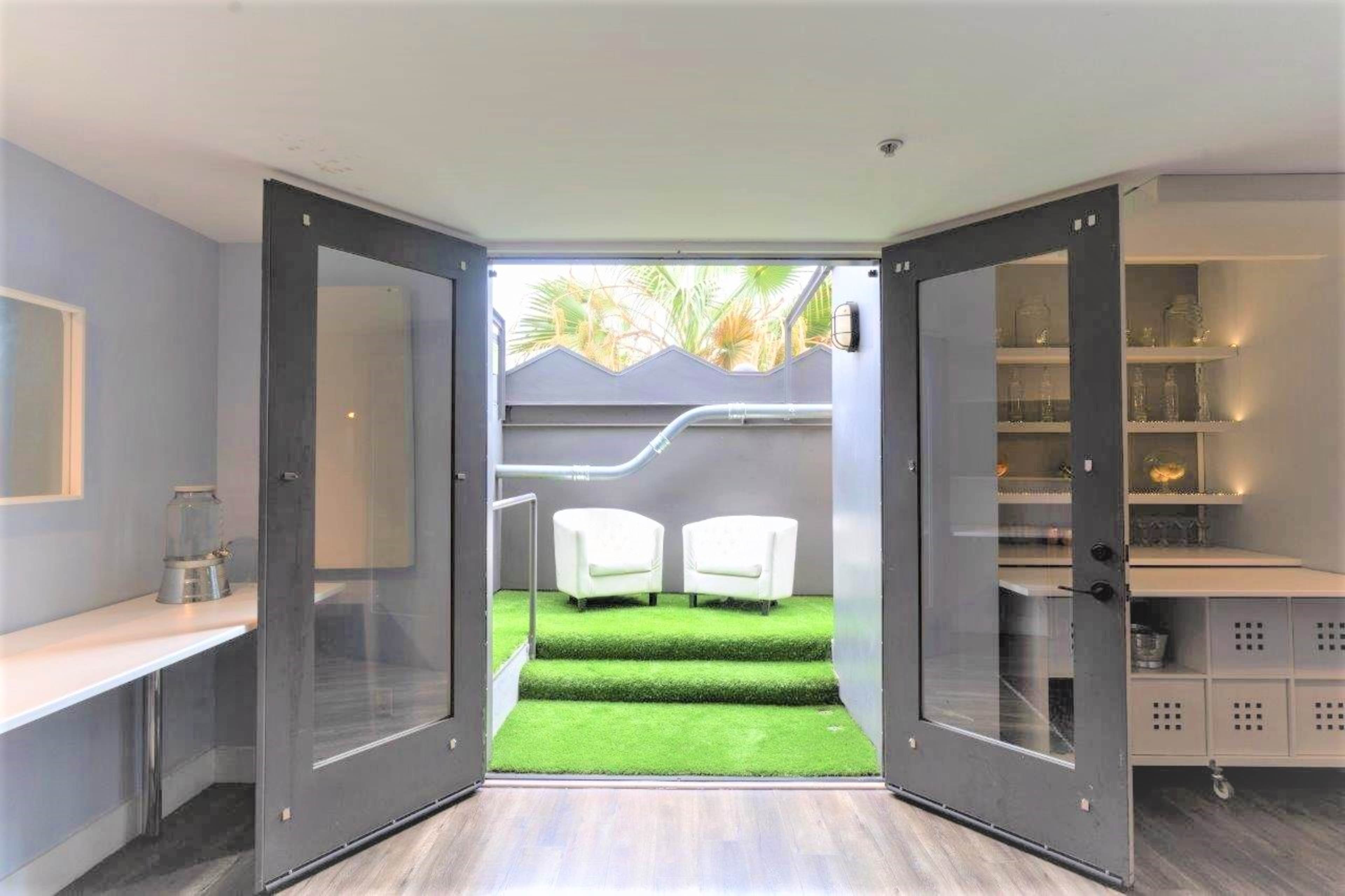 A room with open double doors leading to a small outdoor area featuring two white chairs on artificial grass.