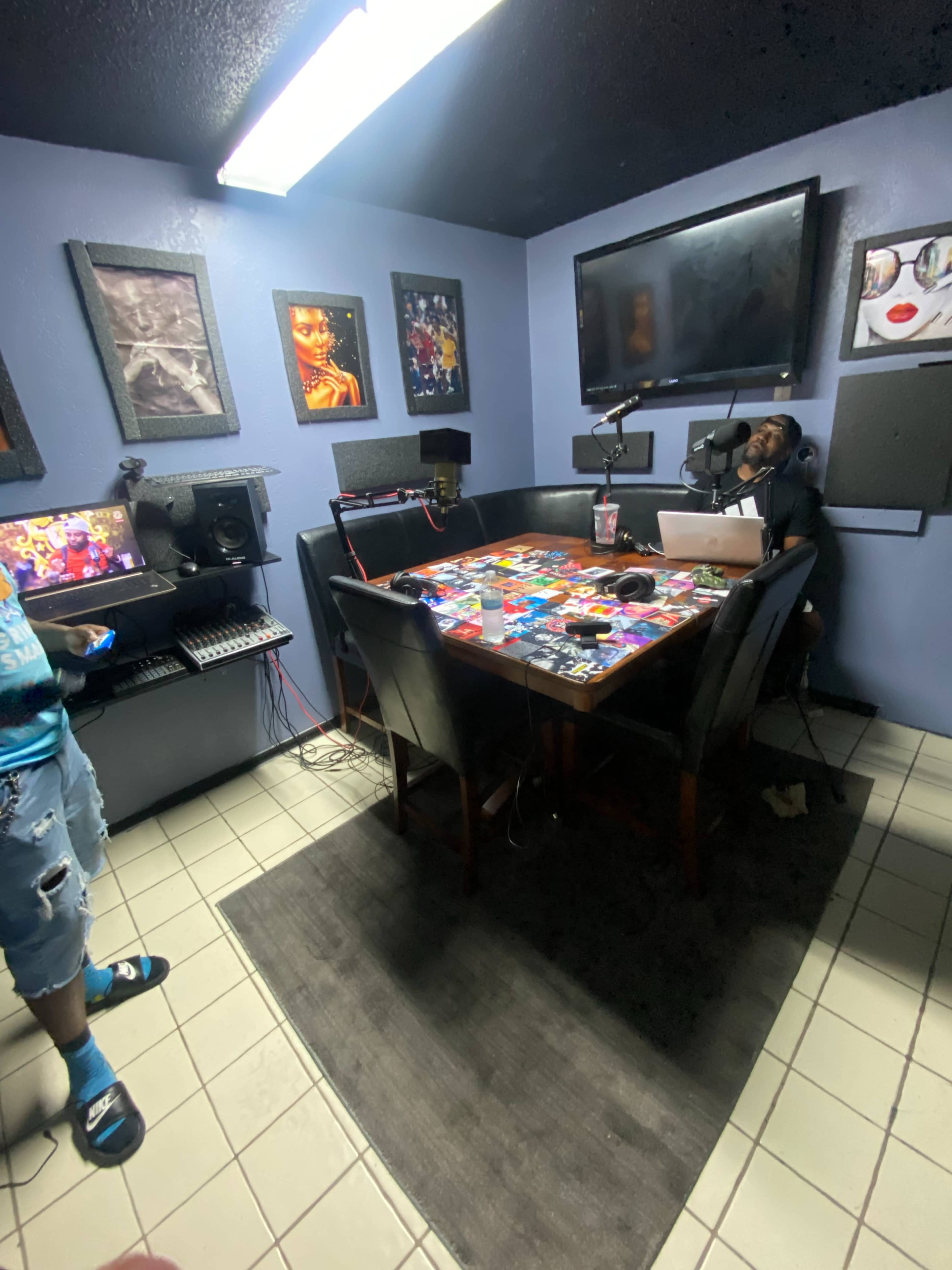 The image shows a small recording studio with a table covered in various items, a computer setup, and artwork on the walls.