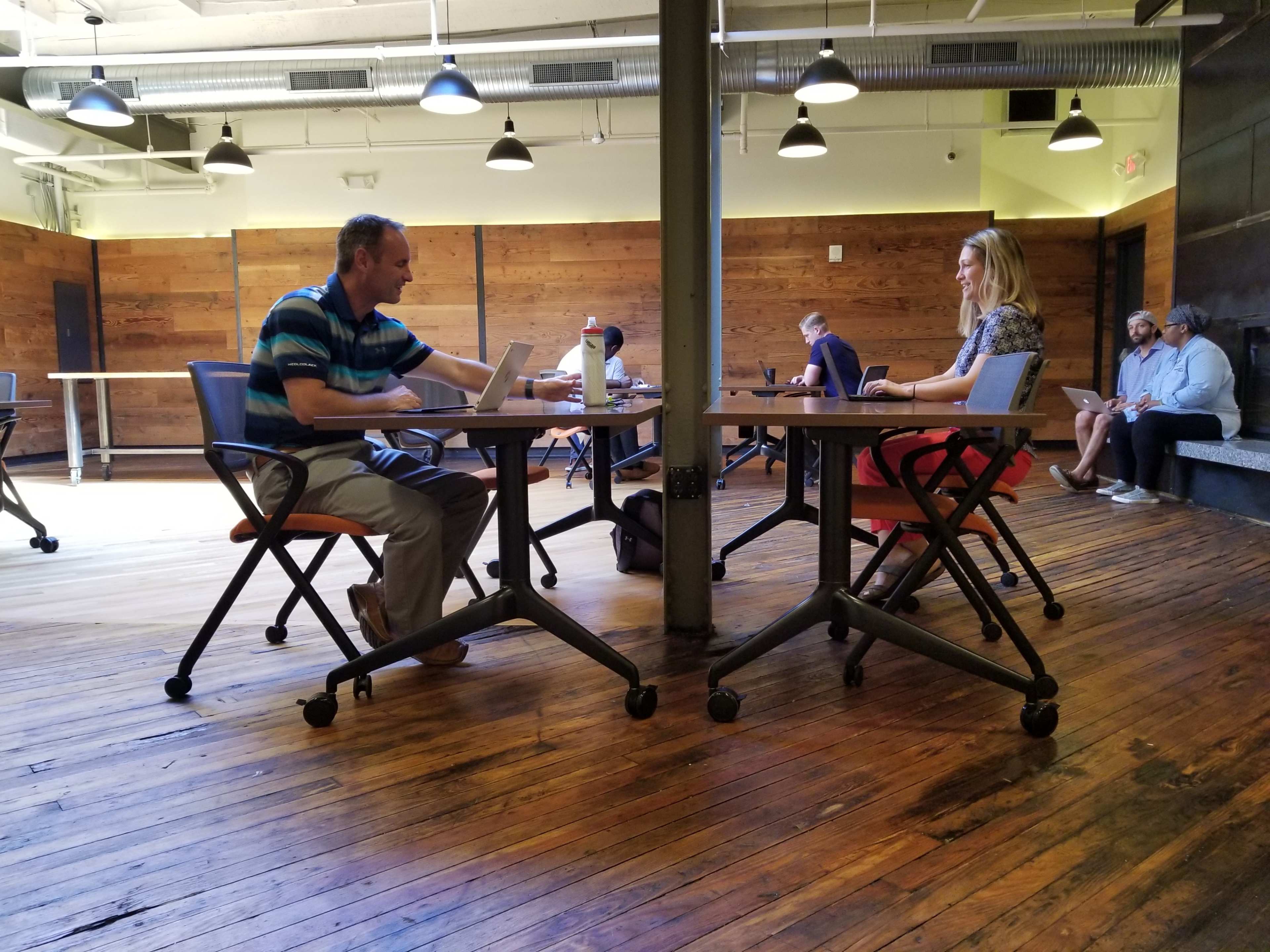 Two people sit at separate tables in a spacious, wood-floored office environment, while others work at their laptops in the background.