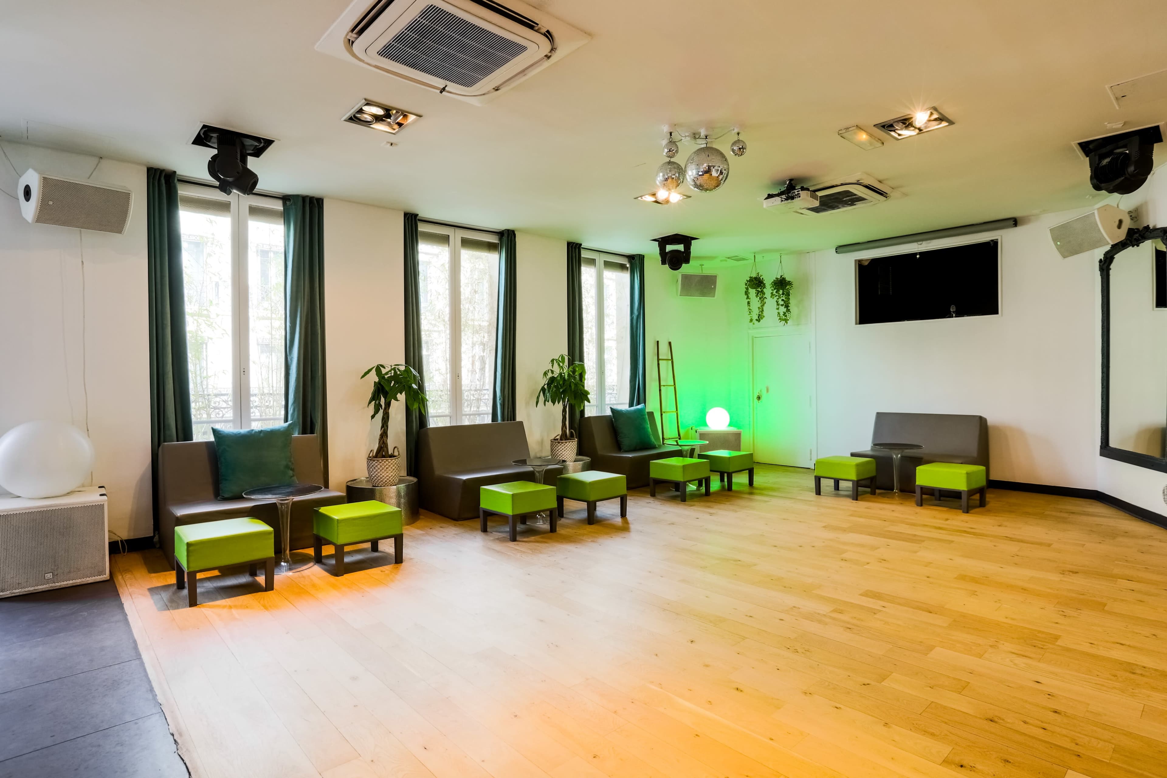 A spacious room with light wooden flooring, green-accented furniture, and large windows allowing natural light in.
