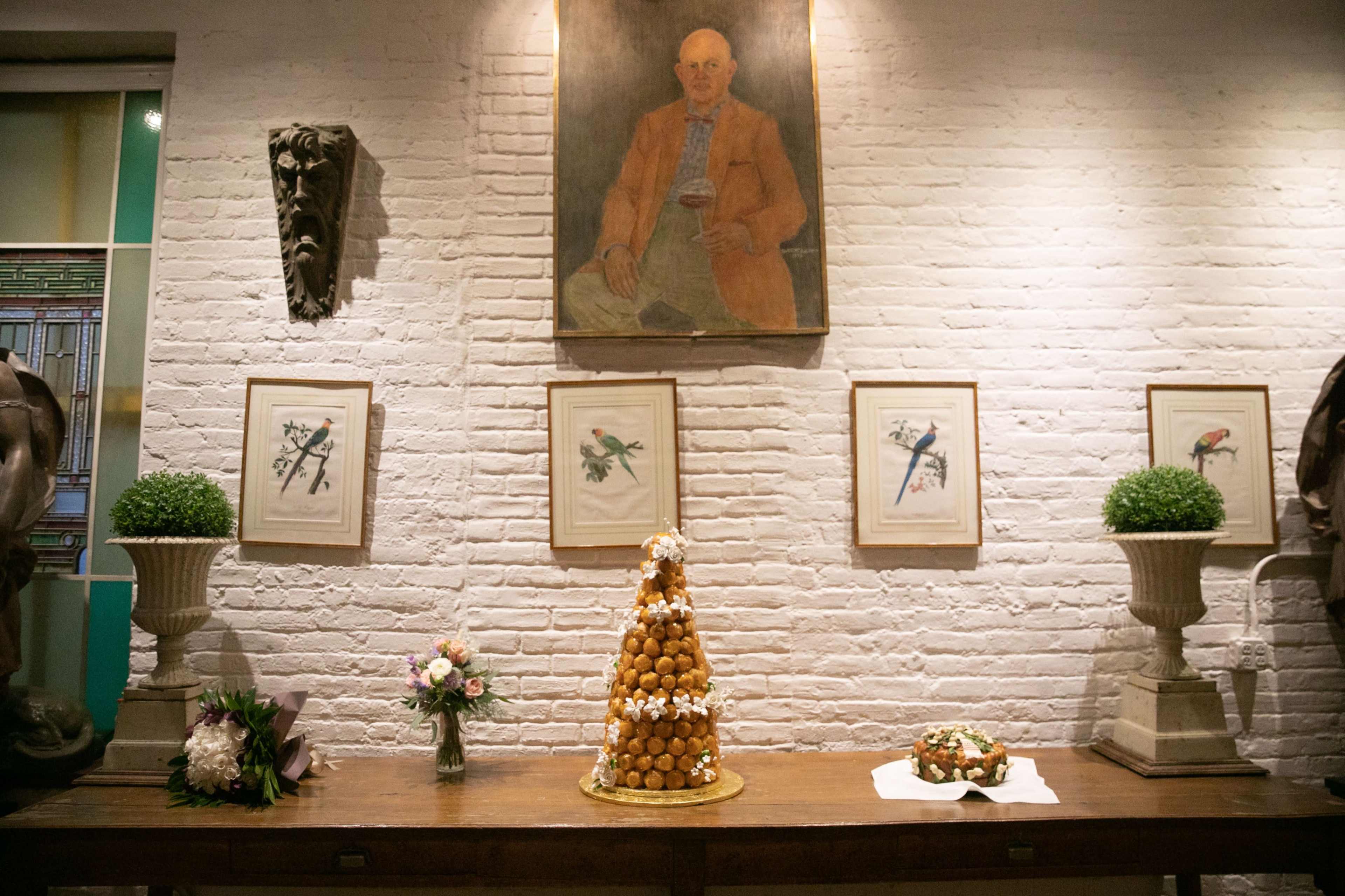 A decorated wall displaying a portrait, framed artworks, and sculptures, with a table featuring a tall dessert made of cream puffs and a floral arrangement.