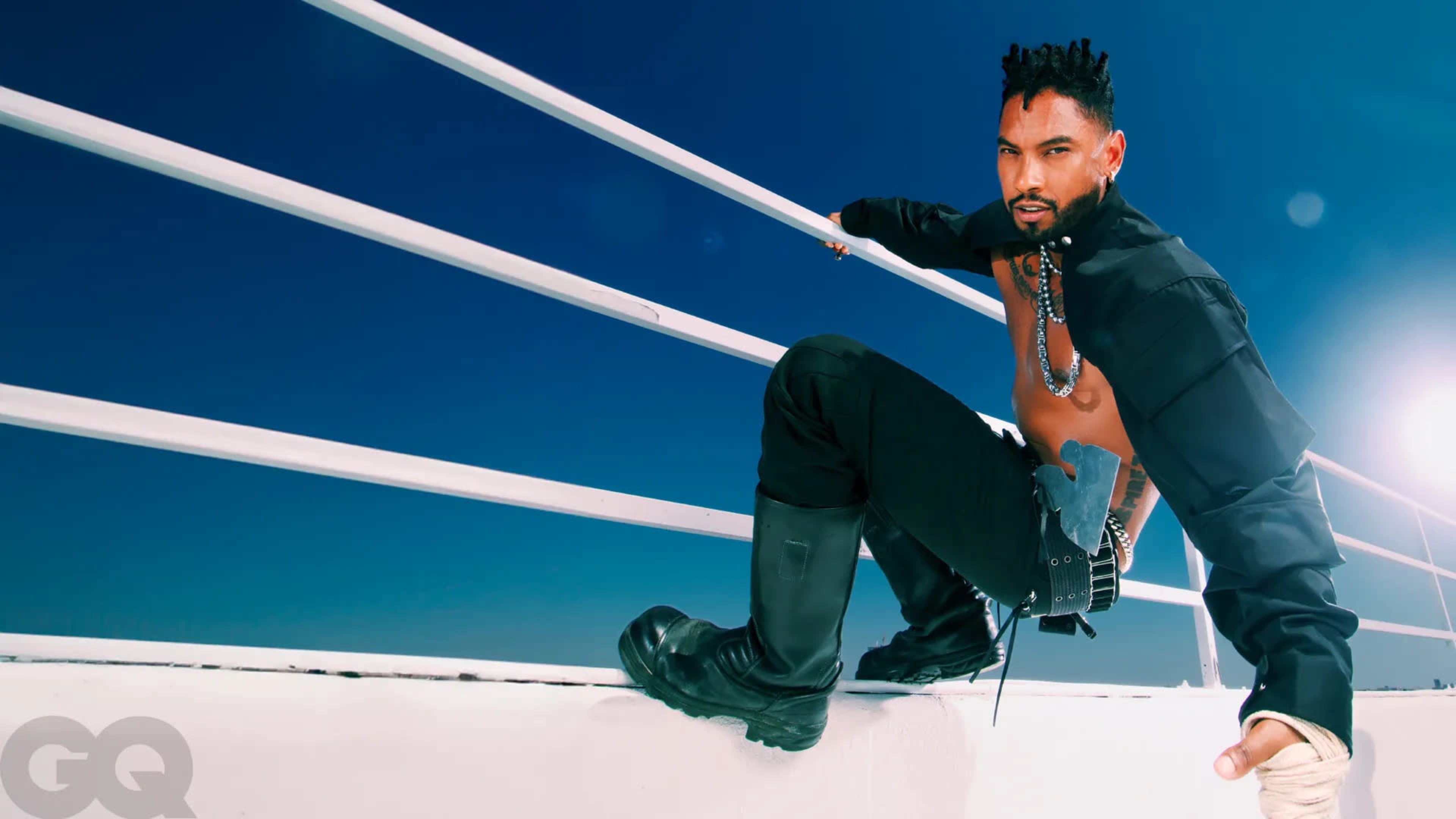A man poses on a railing against a clear blue sky, wearing a cropped jacket and boots.