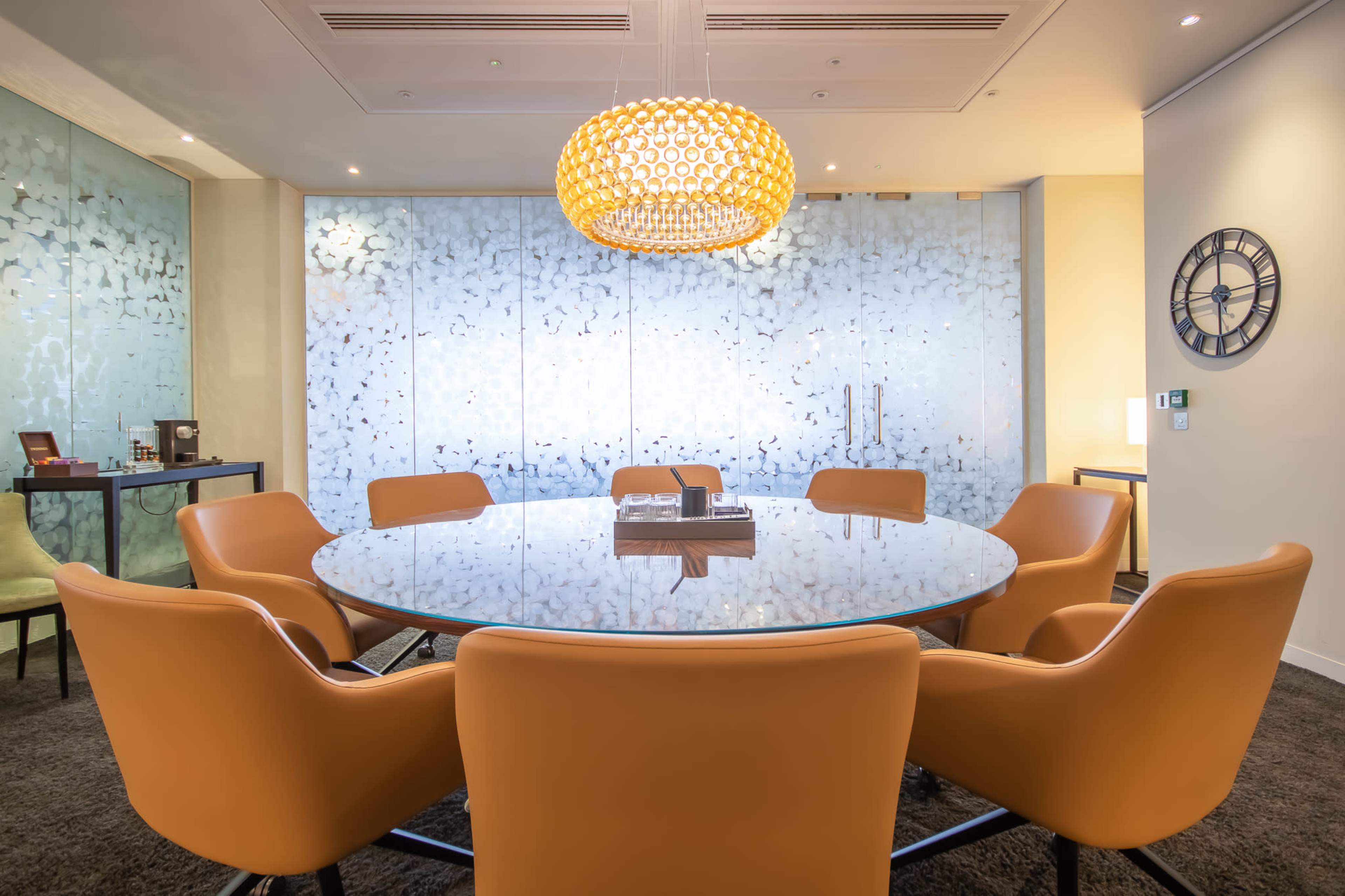 A round glass meeting table surrounded by orange chairs is set in a modern room with a decorative light fixture and a patterned glass wall.