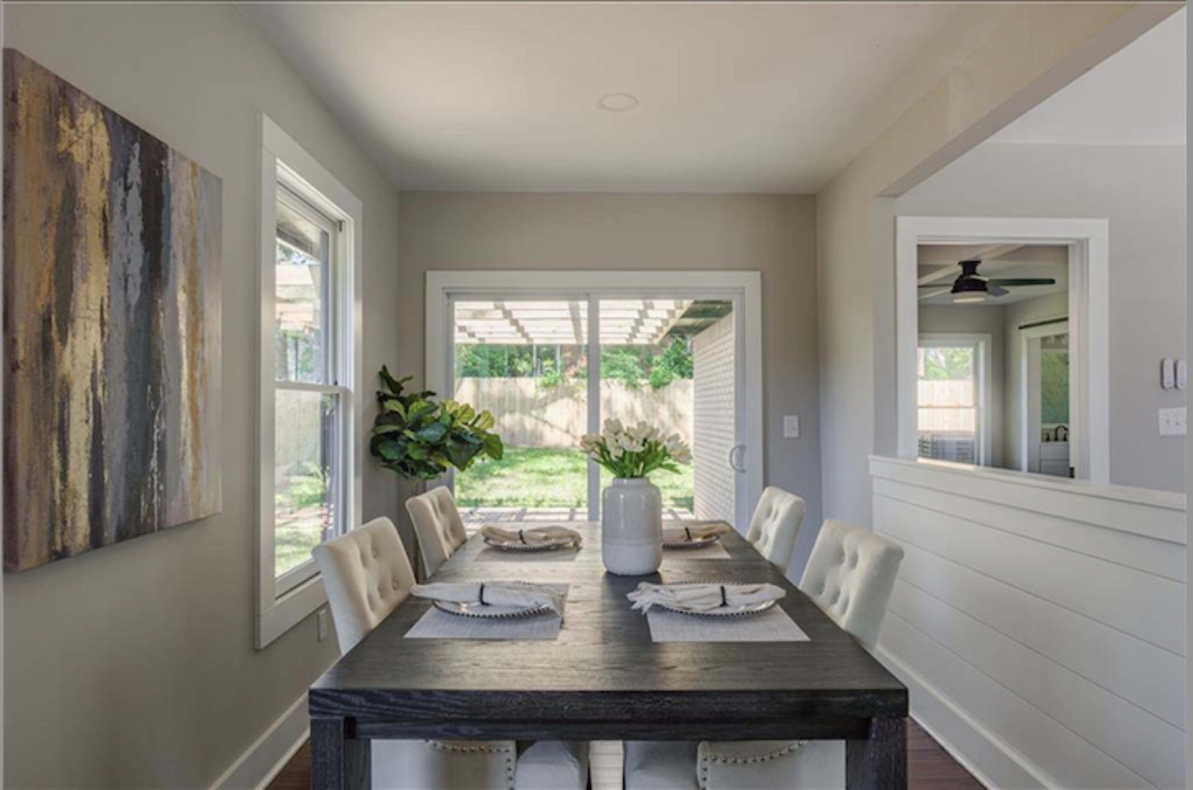 The image shows a dining area with a wooden table set for six, accompanied by chairs, and a view of a backyard through large glass doors.