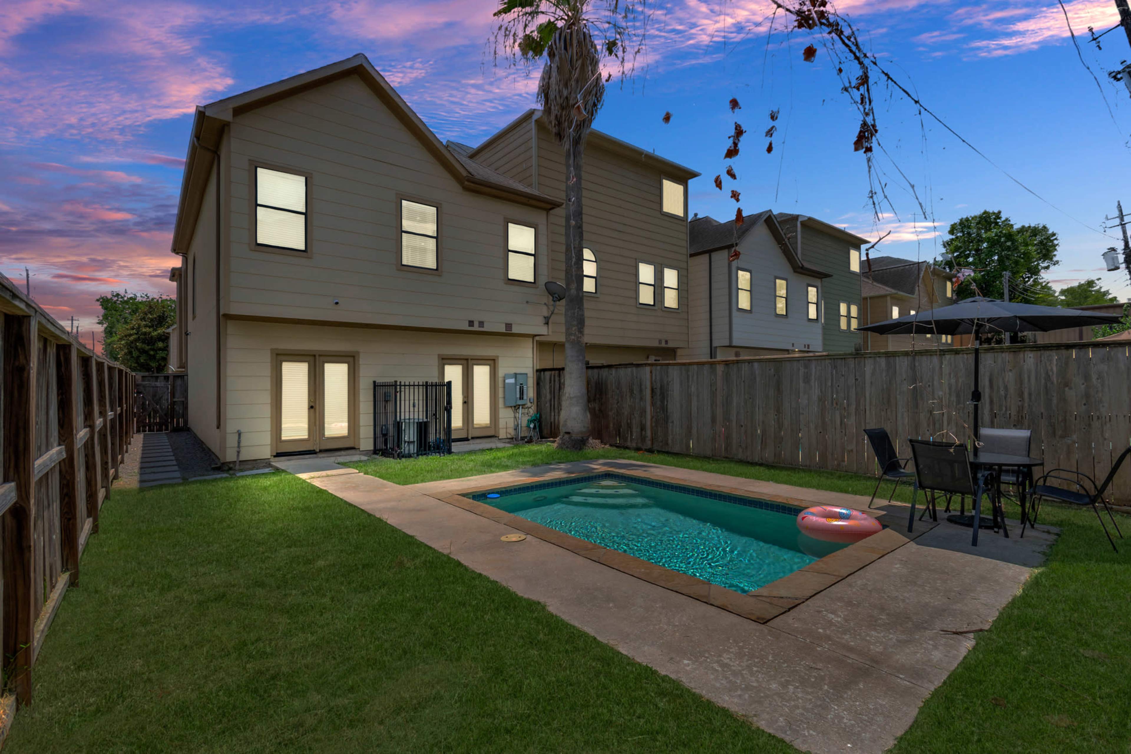 A backyard featuring a swimming pool surrounded by a grassy area, with a two-story house and a wooden fence in the background.