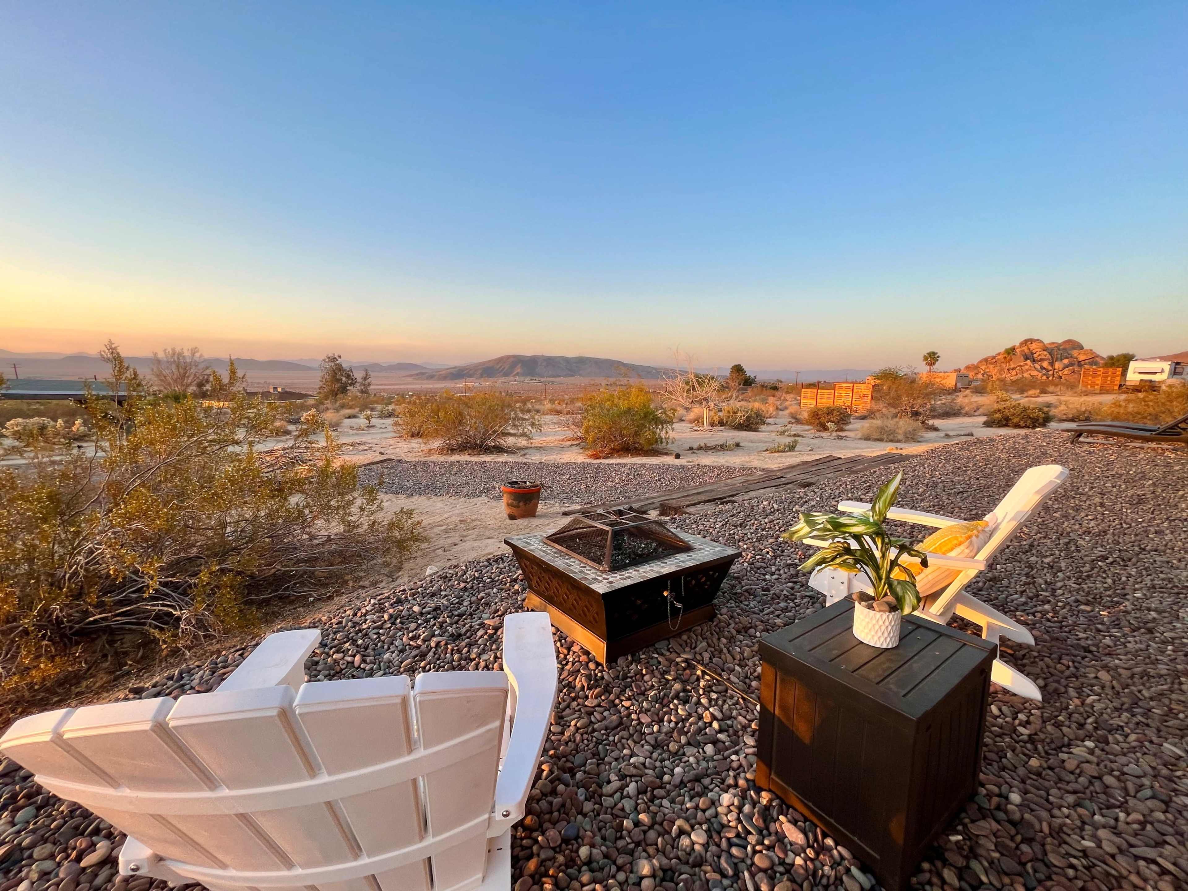 The scene shows a desert landscape at sunset with two white chairs, a black fire pit, and a potted plant on a gravel surface.