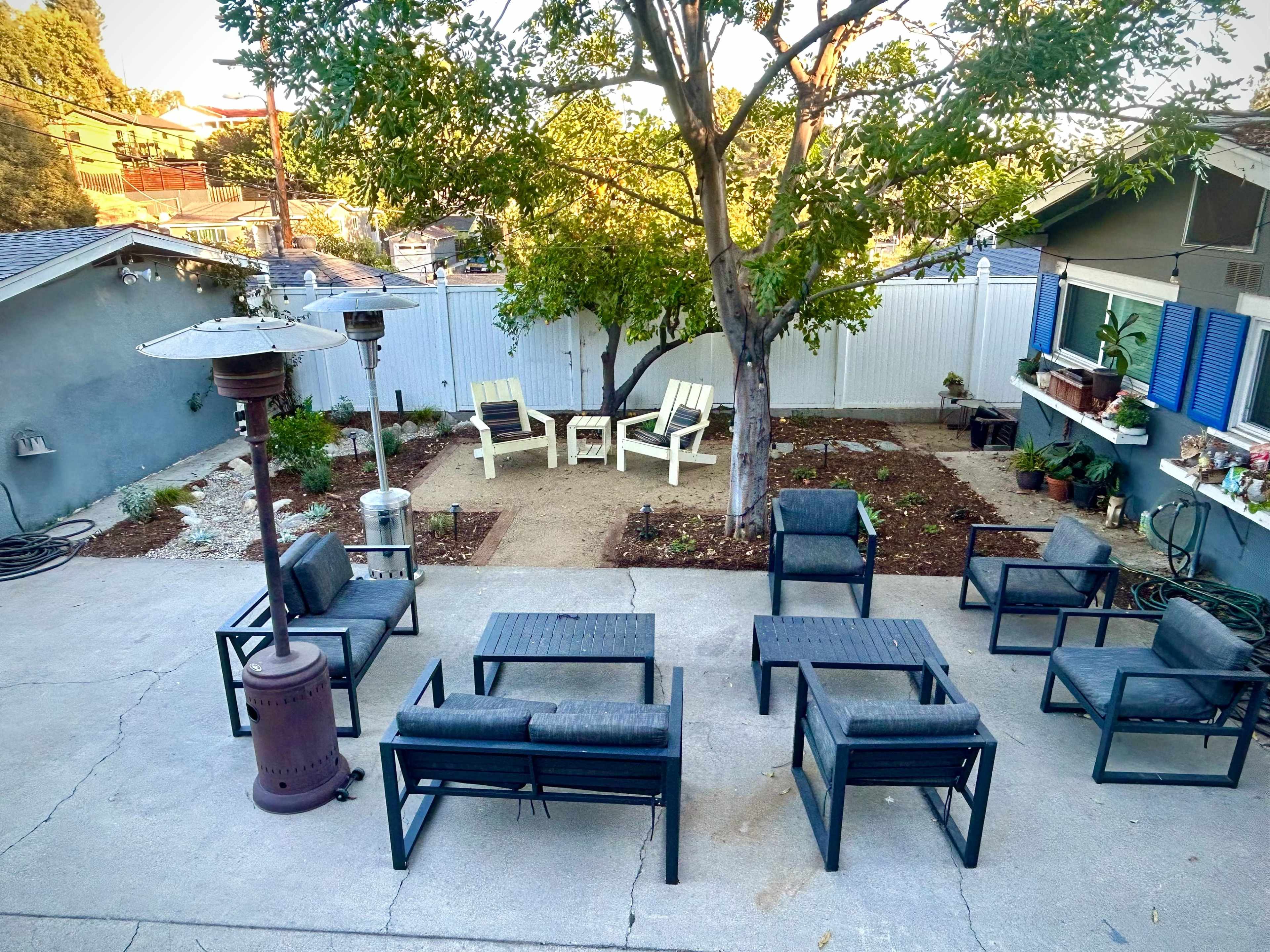 A backyard patio features multiple outdoor seating arrangements, a heater, and a tree centered in the garden area.