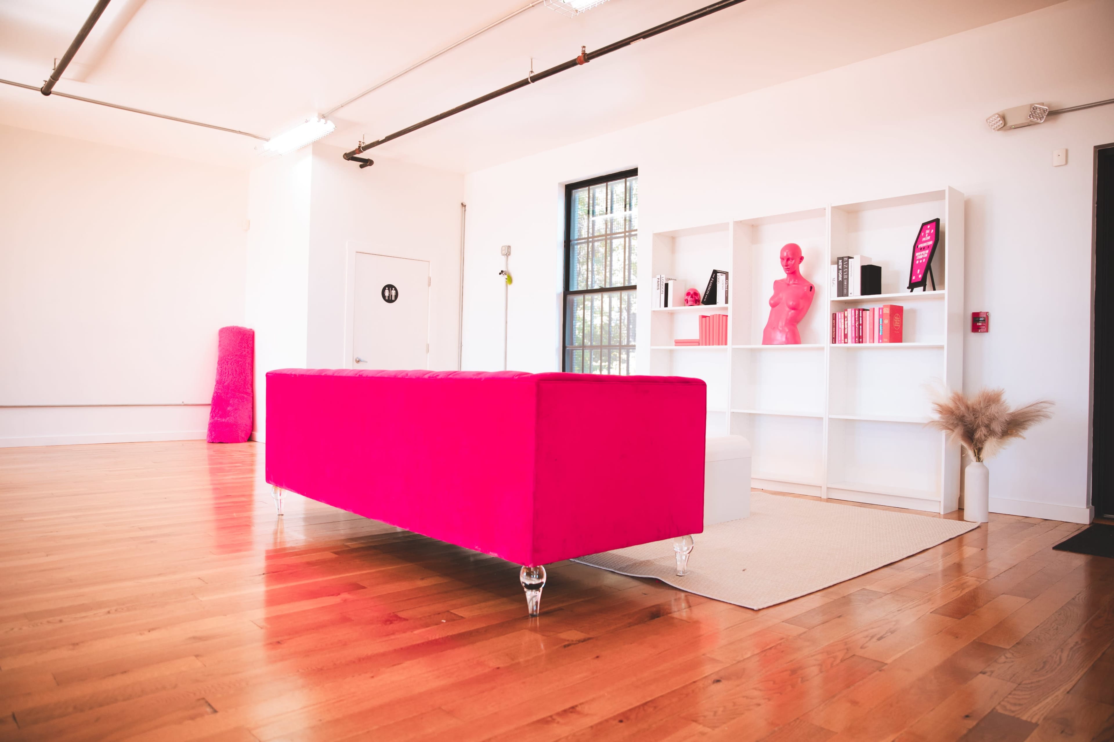 A bright pink sofa is positioned in a spacious room with light wooden flooring, accompanied by a white bookshelf containing various items, including a pink sculpture.