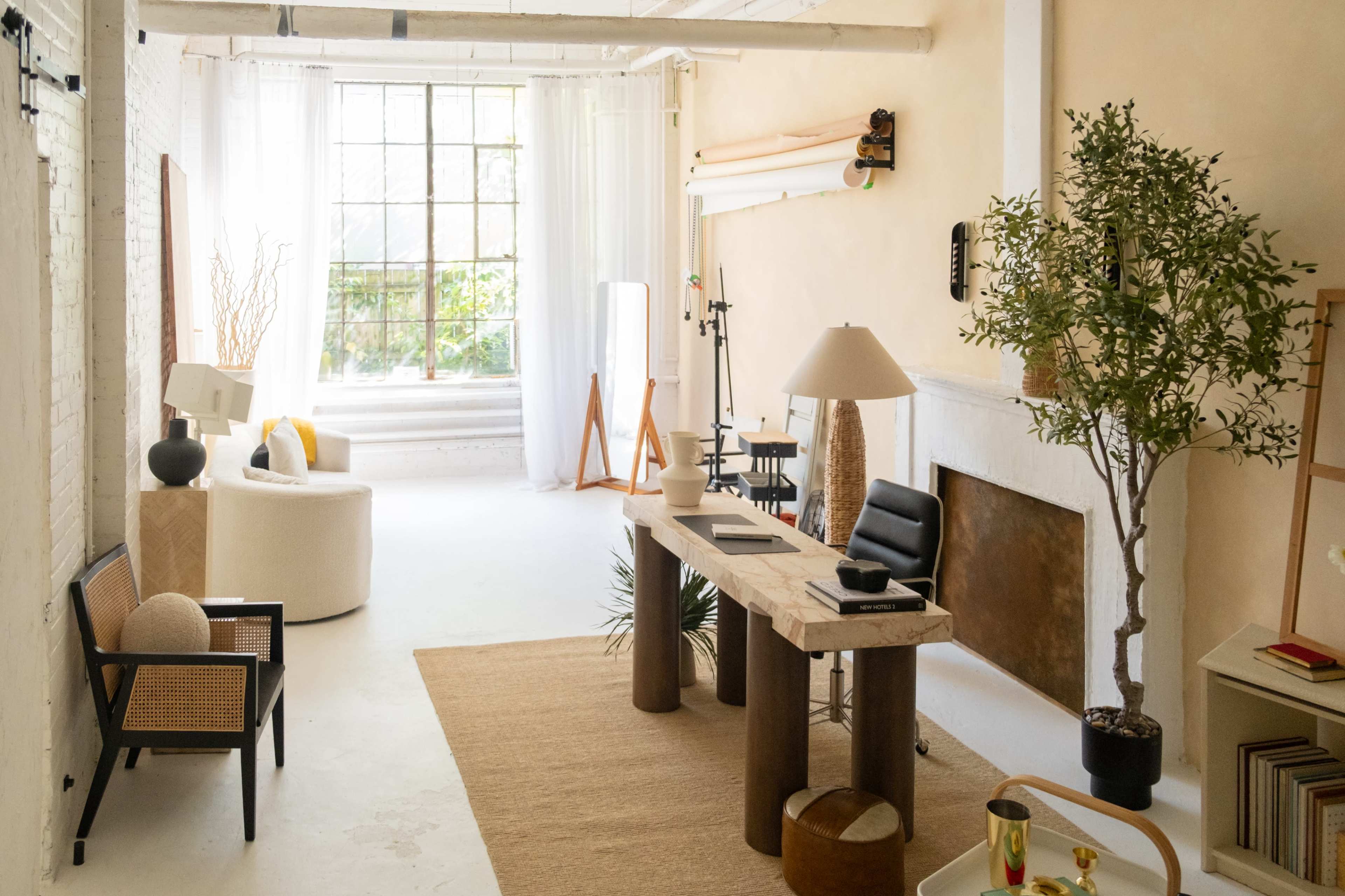 The image shows a spacious, well-lit room featuring a wooden desk, a modern chair, various plants, and minimalist decor elements.