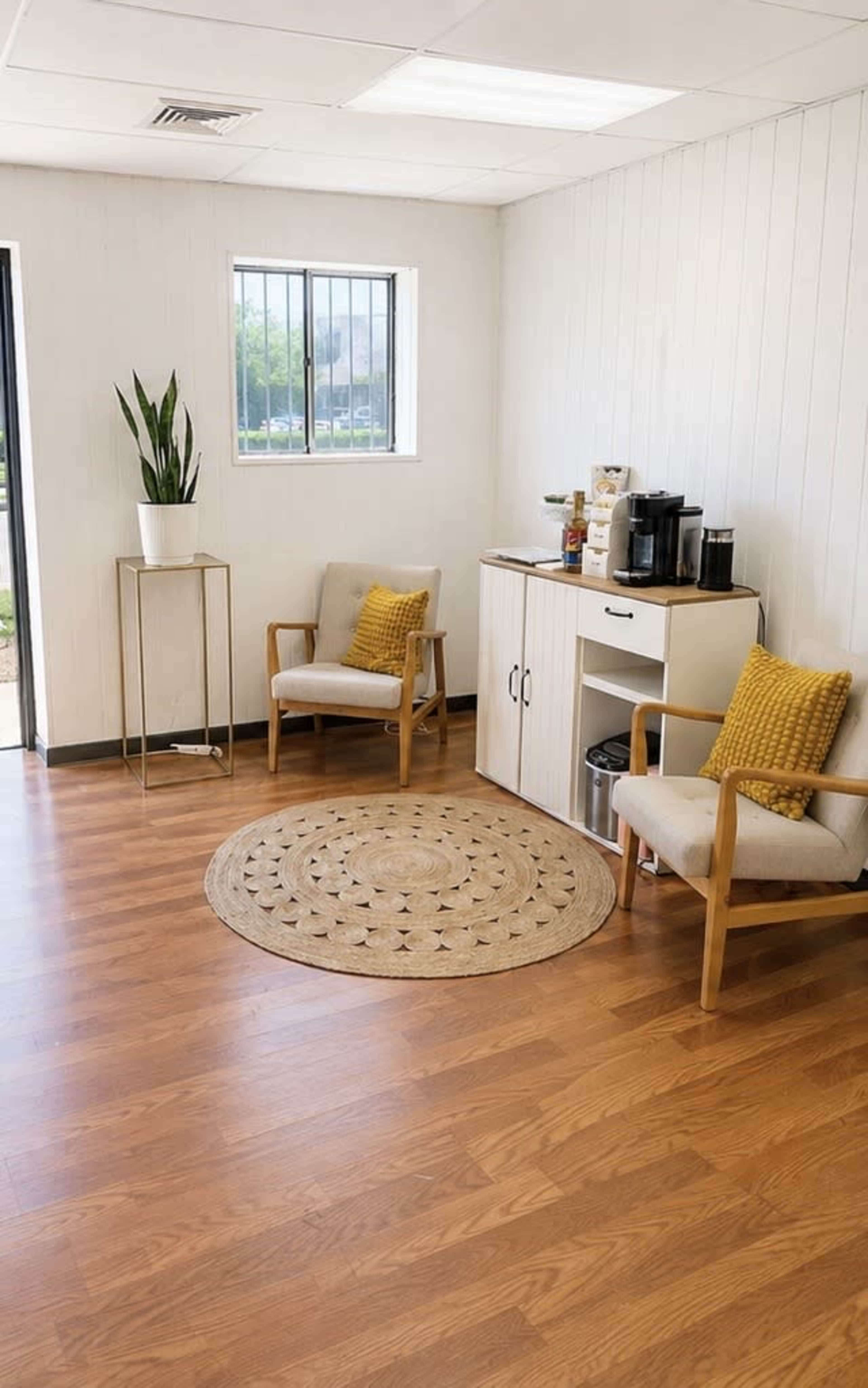 The image shows a small breakroom with two chairs, a round rug, a cabinet with coffee supplies, and a plant in the corner.