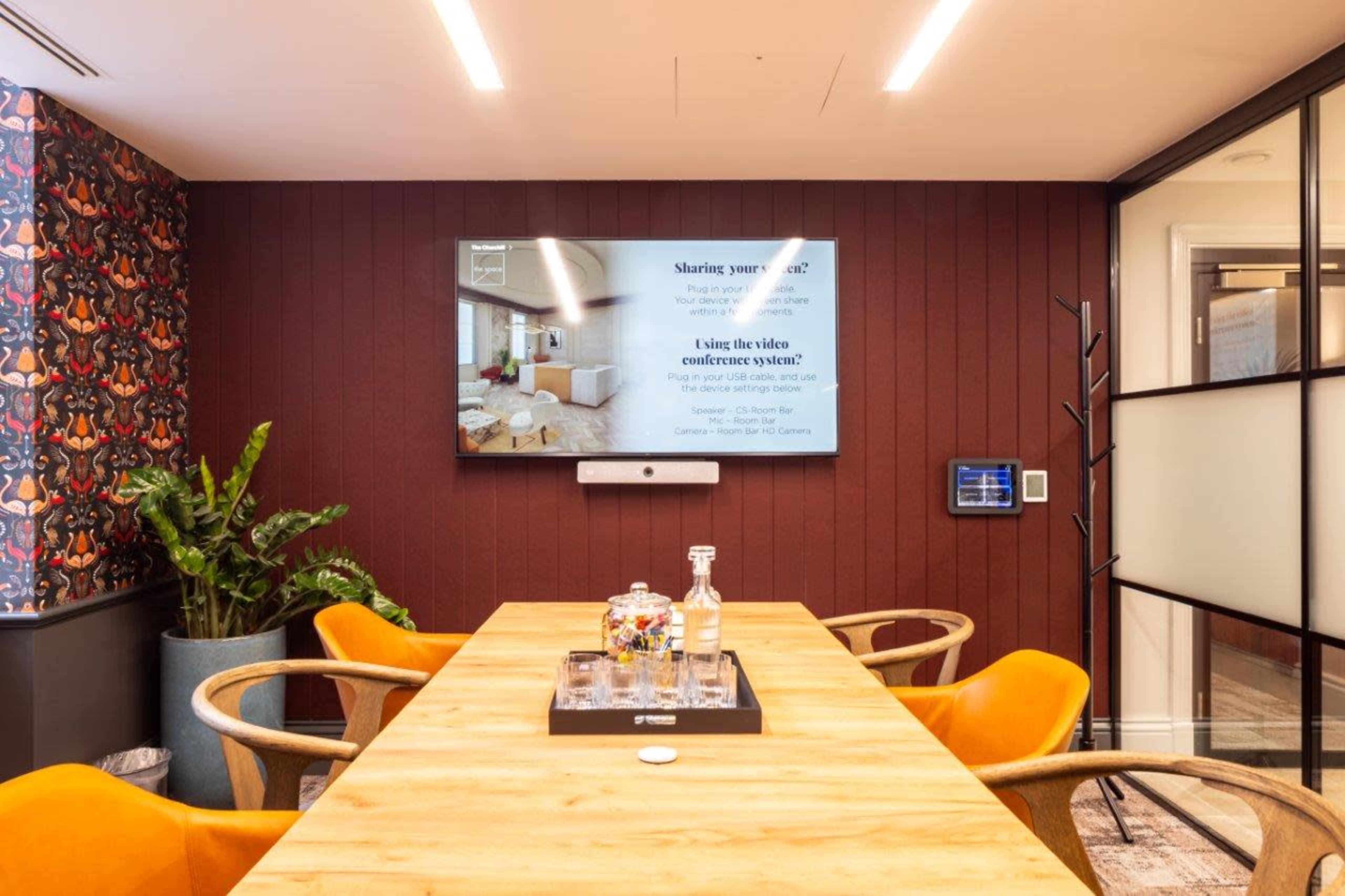 A modern conference room is equipped with a large screen displaying information, a wooden table surrounded by orange chairs, and a plant in the corner.