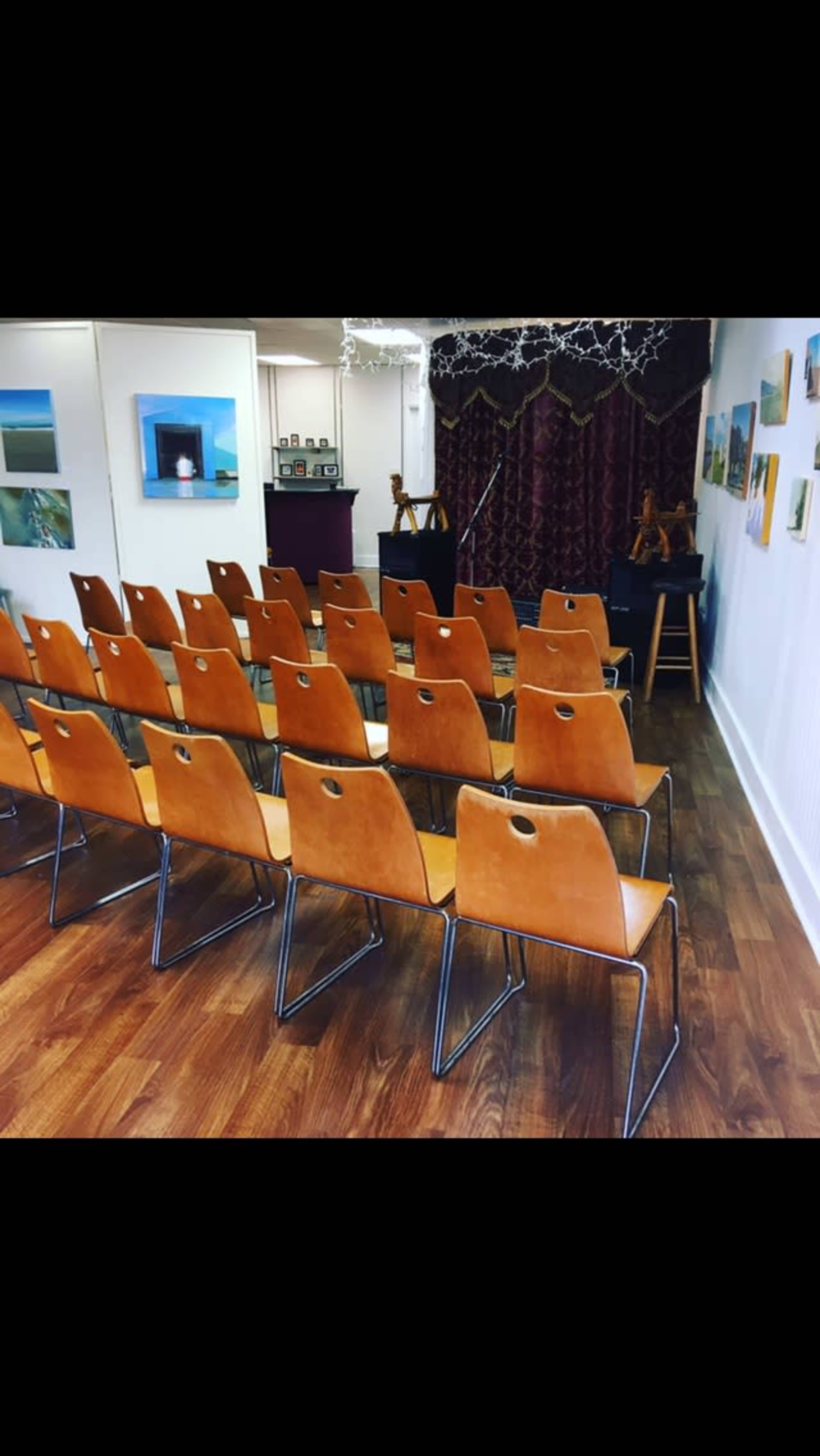 The image shows a room set up for an event with rows of orange chairs facing a stage draped in red, along with artwork displayed on the walls.