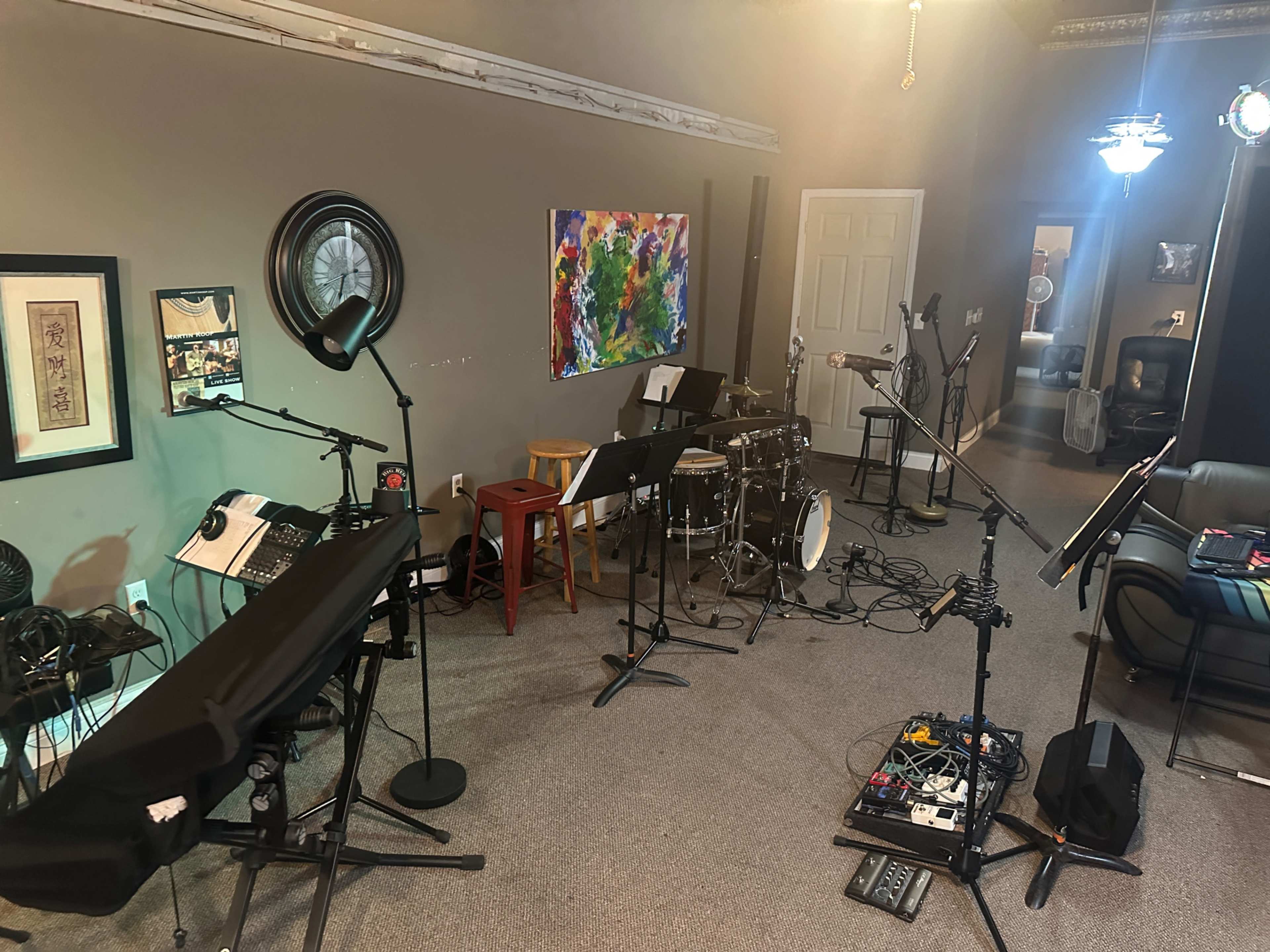 The image shows a music studio setup with various instruments including a keyboard, drums, and microphones, alongside a colorful abstract painting on the wall.