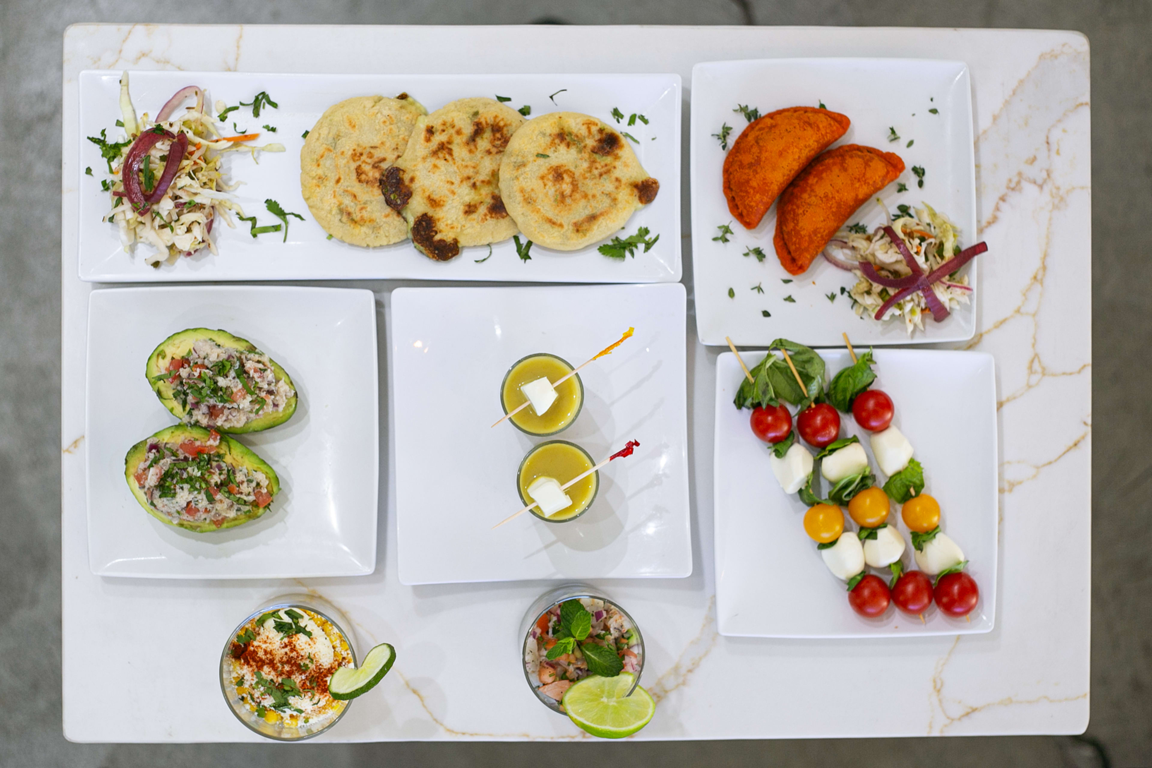 The image showcases a variety of colorful dishes, including stuffed avocados, arepas, empanadas, salads, and skewered vegetables, arranged on white square plates on a marble table.