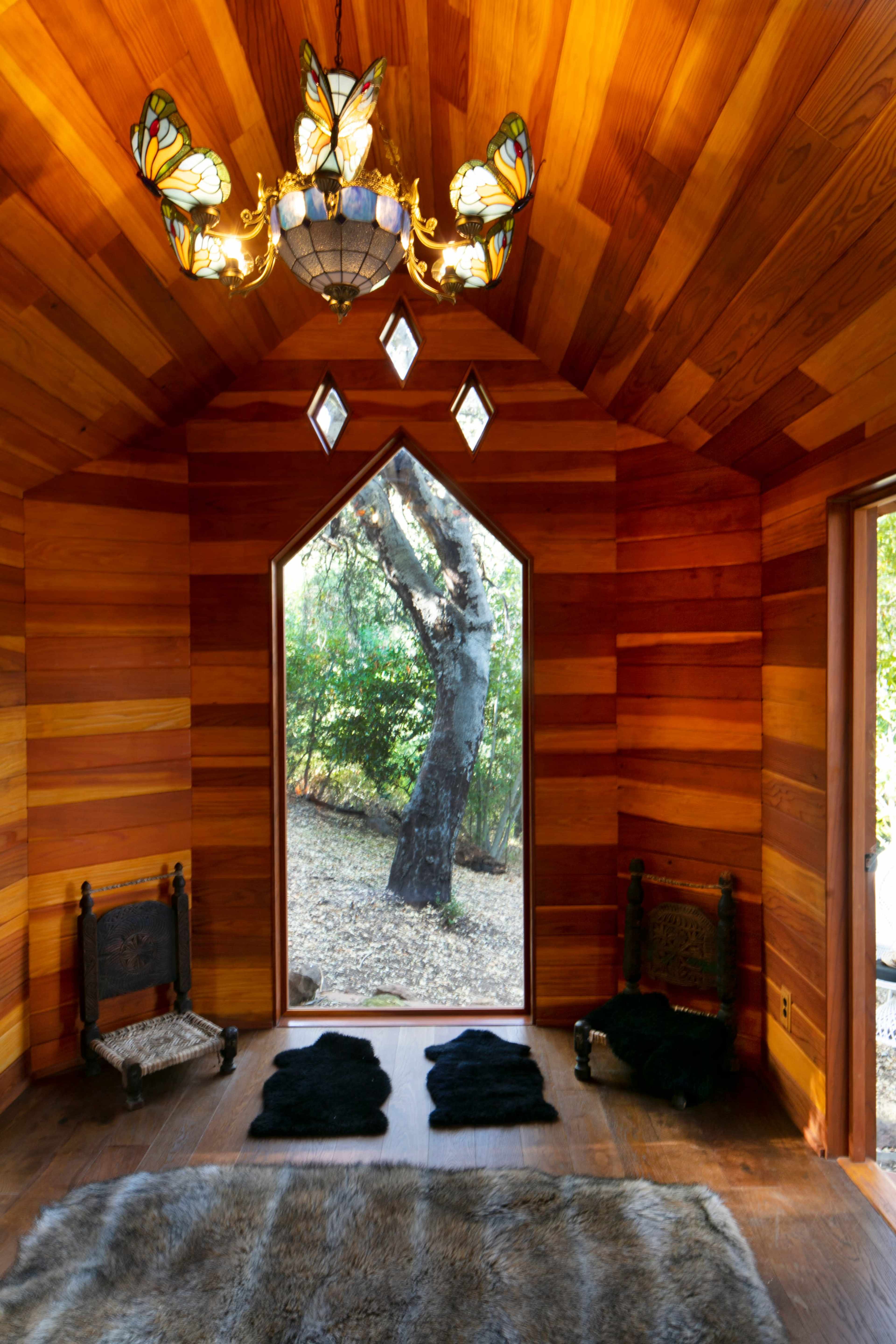 The interior of a wooden room features a butterfly-themed chandelier and two ornate chairs flanking a large window that offers a view of a tree outside.
