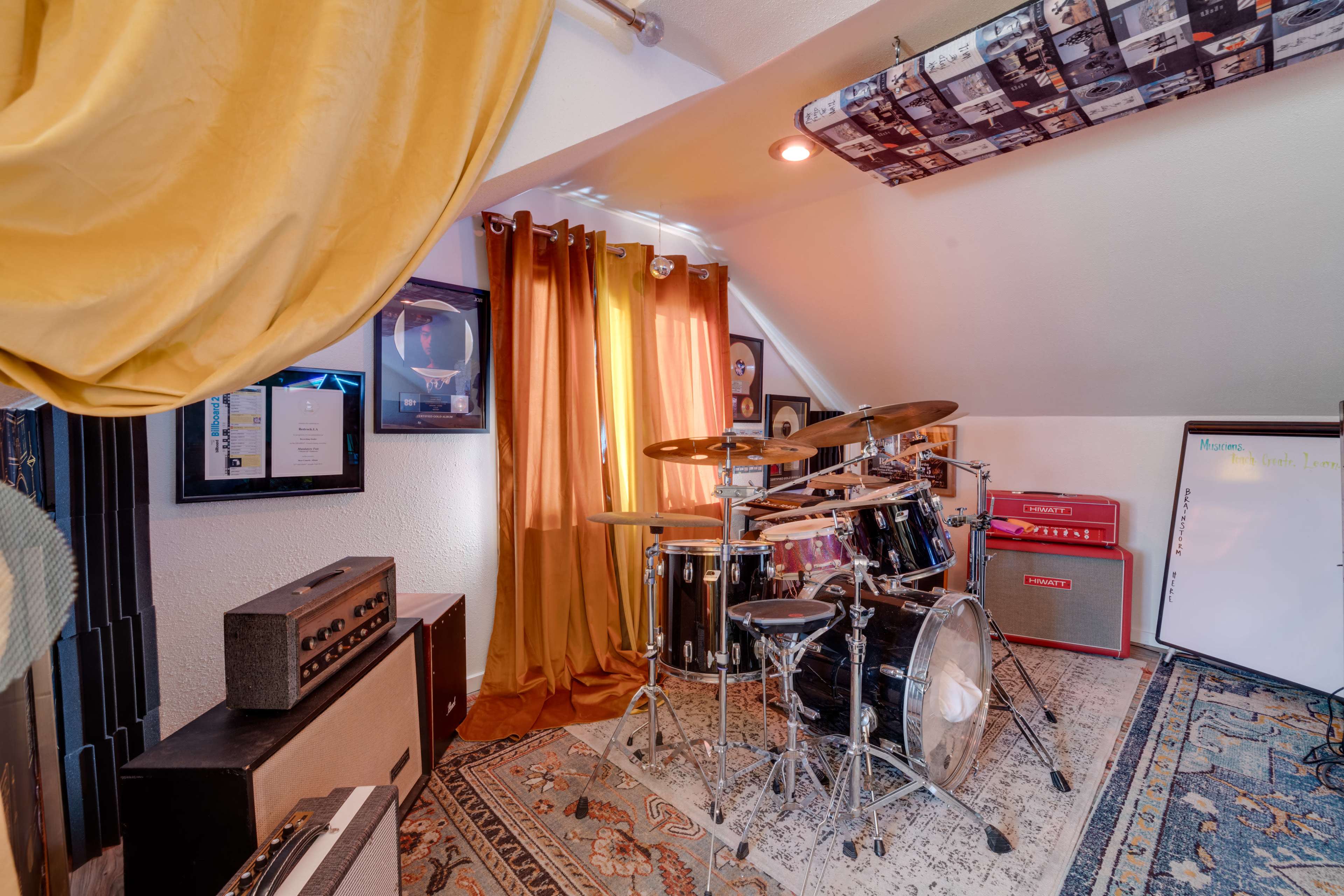 The image shows a music rehearsal room featuring a drum set, amplifiers, and a whiteboard, with warm-colored curtains and a patterned rug.