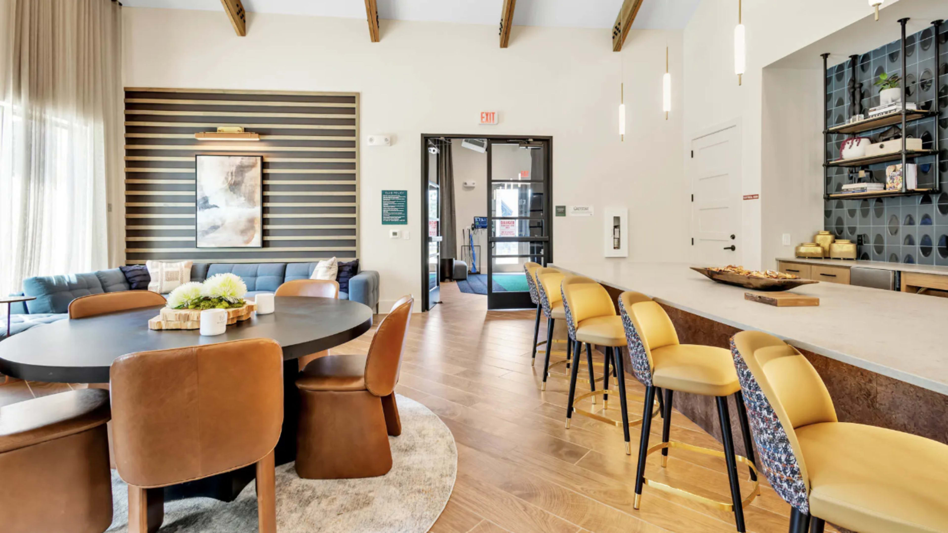 The image shows a modern common area featuring a dining table with upholstered chairs, a bar countertop with stools, and a lounge area with a sofa, all within a well-lit space that has vaulted ceilings.