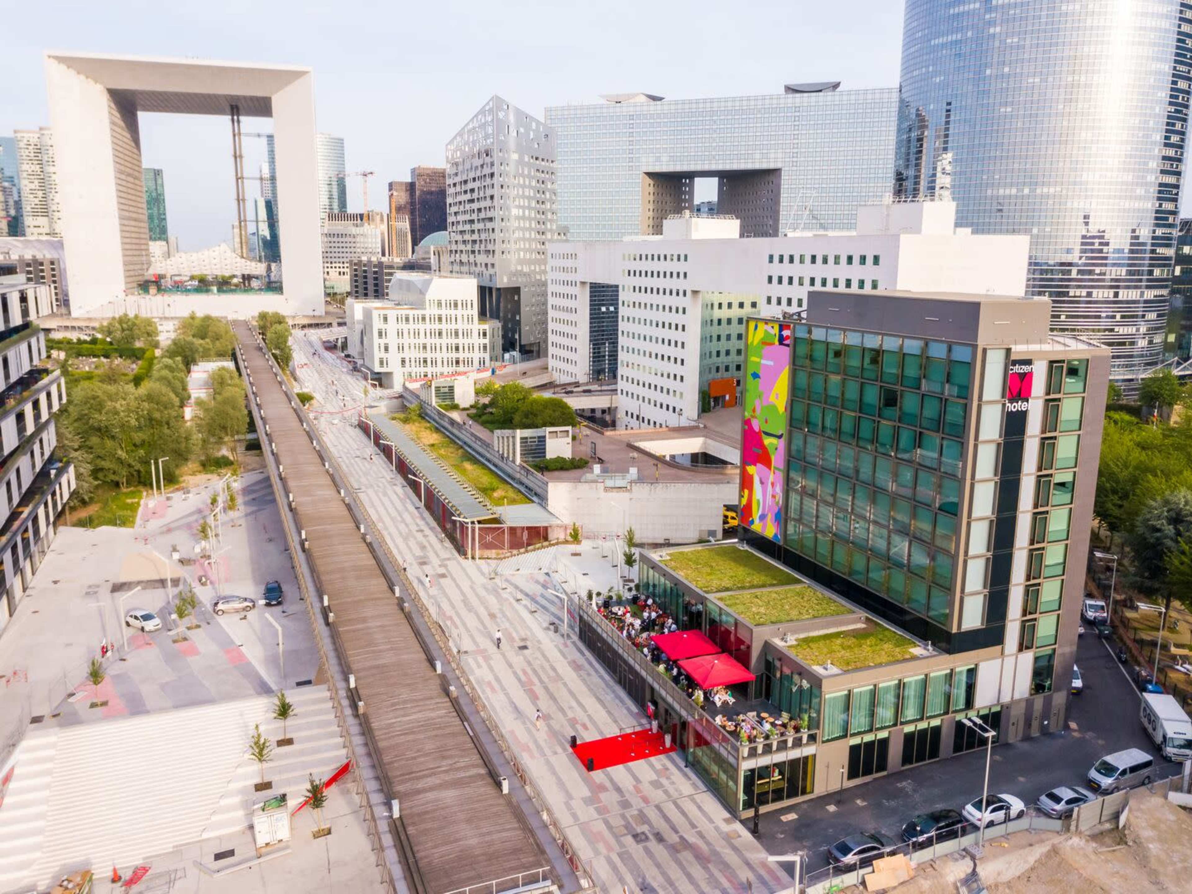 A modern urban landscape featuring a vibrant hotel with colorful artwork, adjacent to a pedestrian walkway and contemporary buildings in La Défense, Paris.