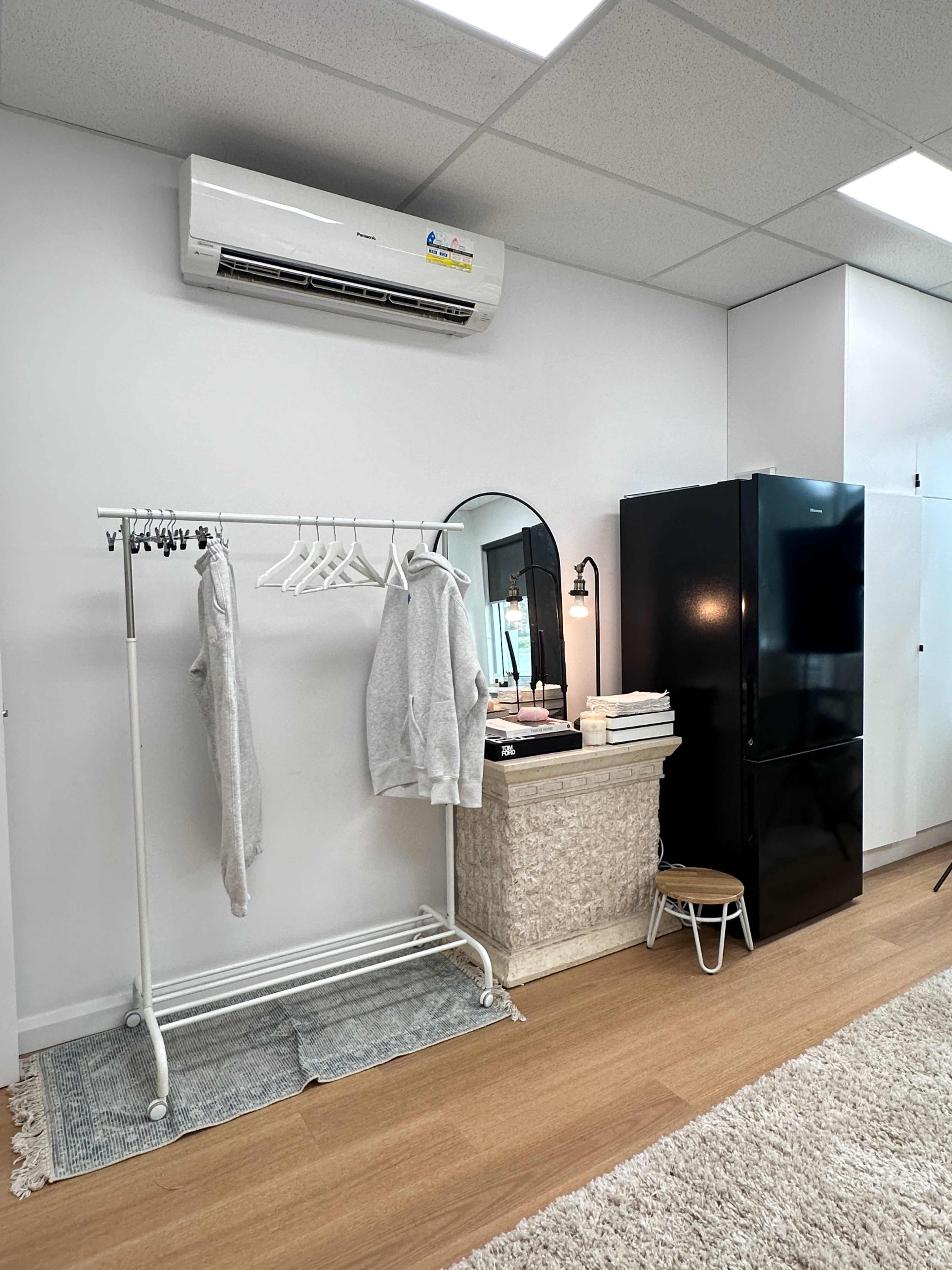 The image shows a small room featuring a clothing rack with gray sweatshirts, a mirror, a black refrigerator, and a white storage unit against a light-colored wall.