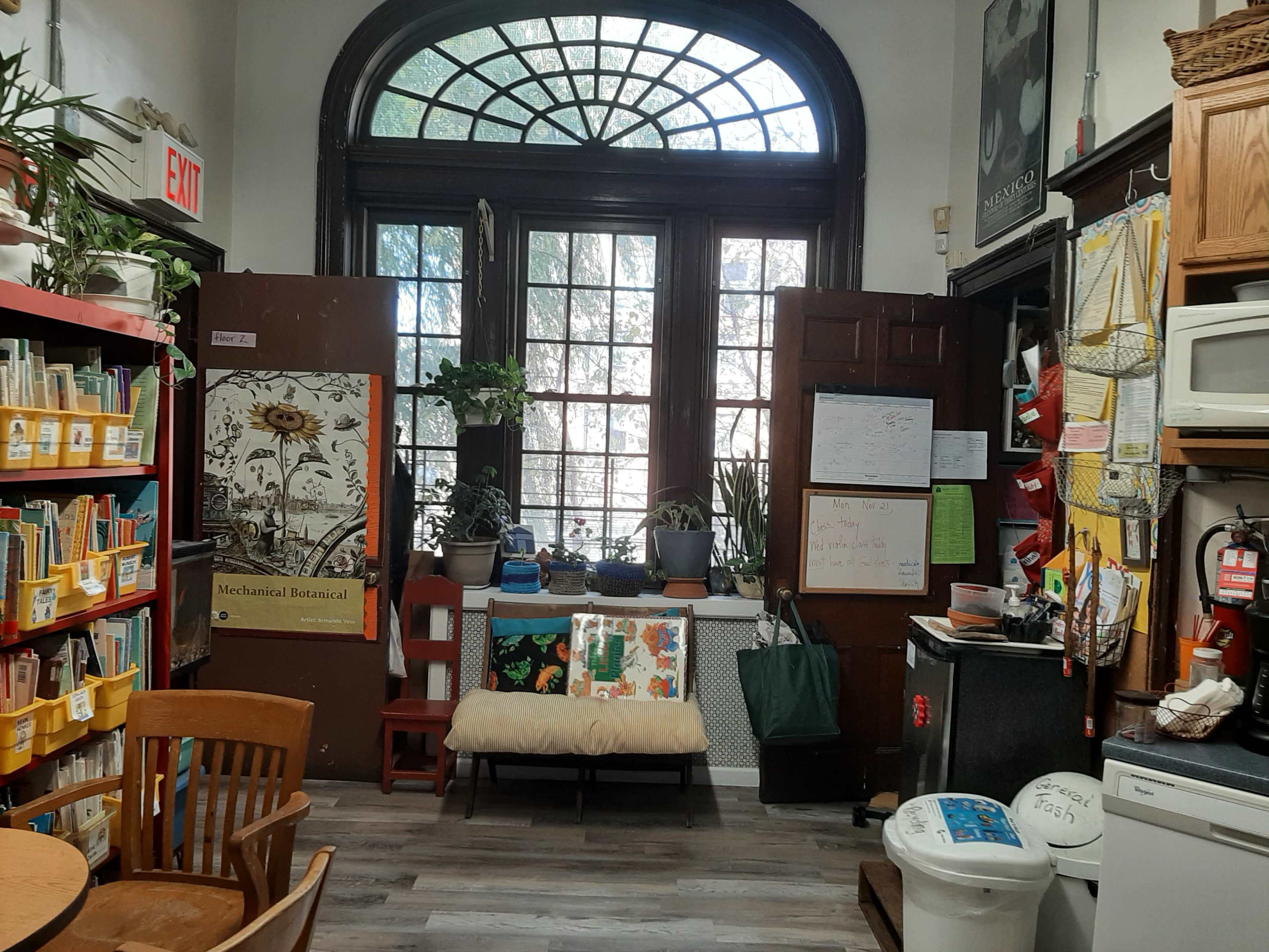 An interior of a cozy room with bookshelves, a bench, kitchen appliances, and large windows that allow light to stream in.