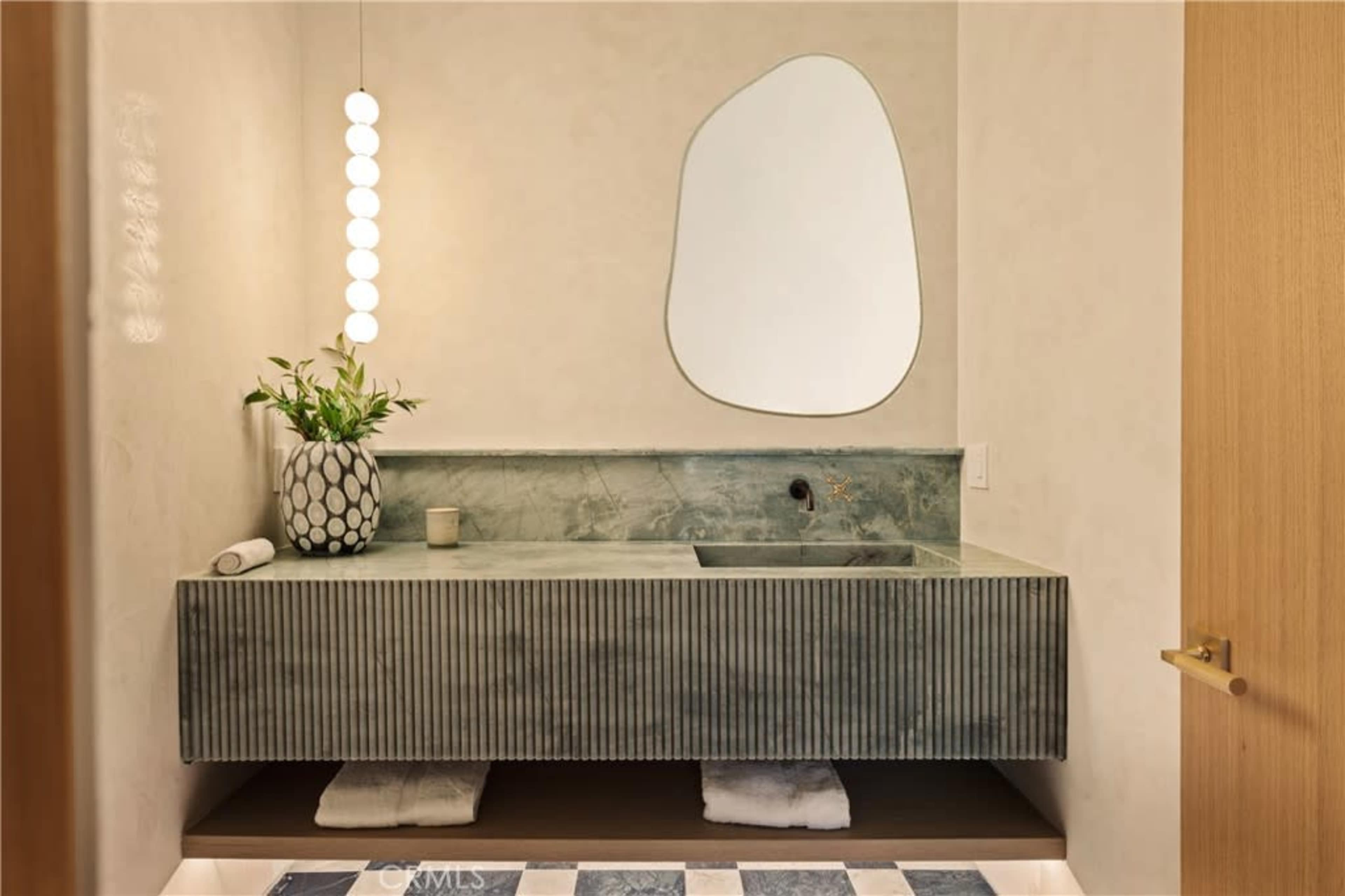 A modern bathroom features a green marble sink countertop with a unique mirror above and a small plant beside it, situated against a neutral wall.