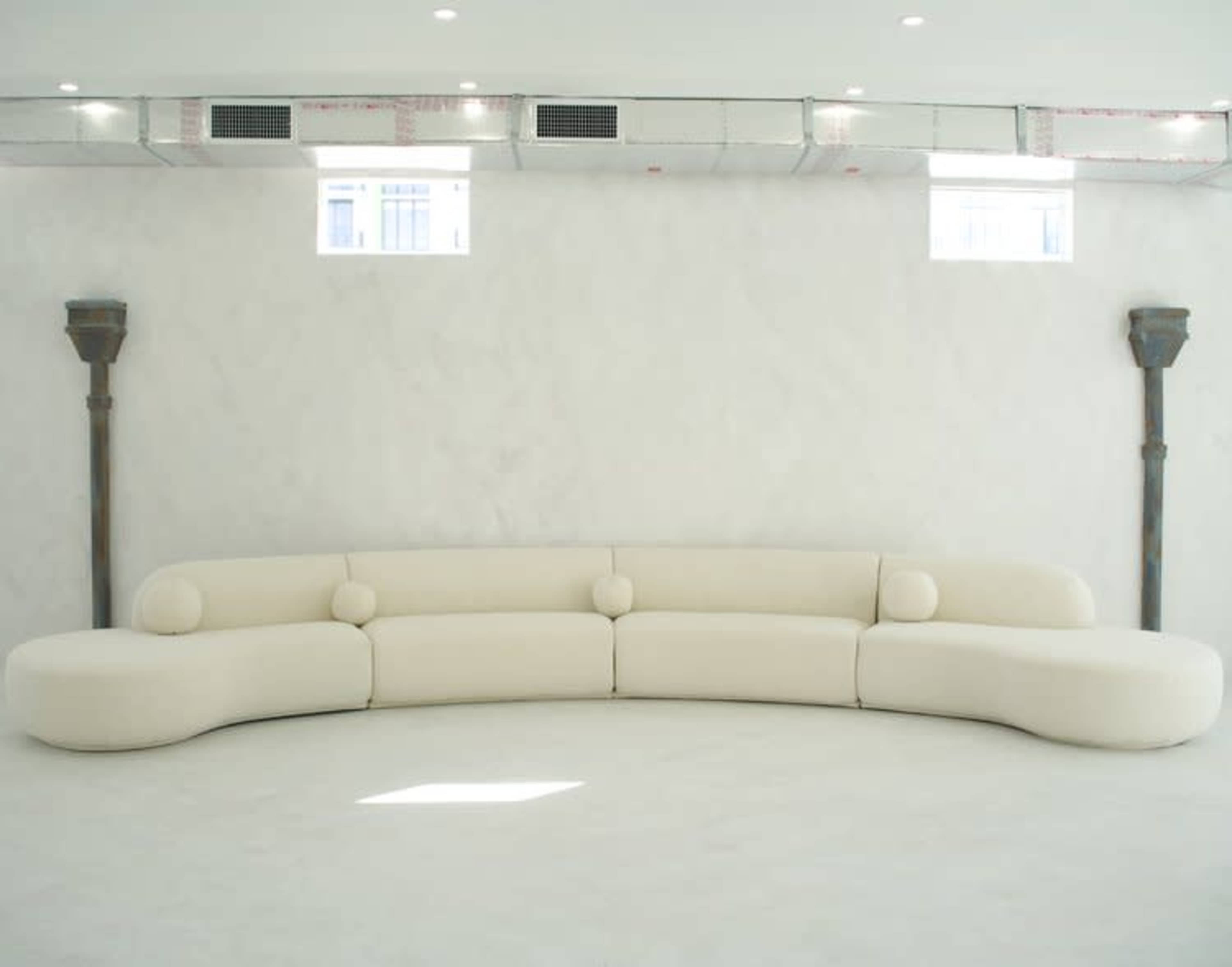 A curved white sofa is positioned against a smooth, light-colored wall with two small windows and exposed ventilation ducts above.