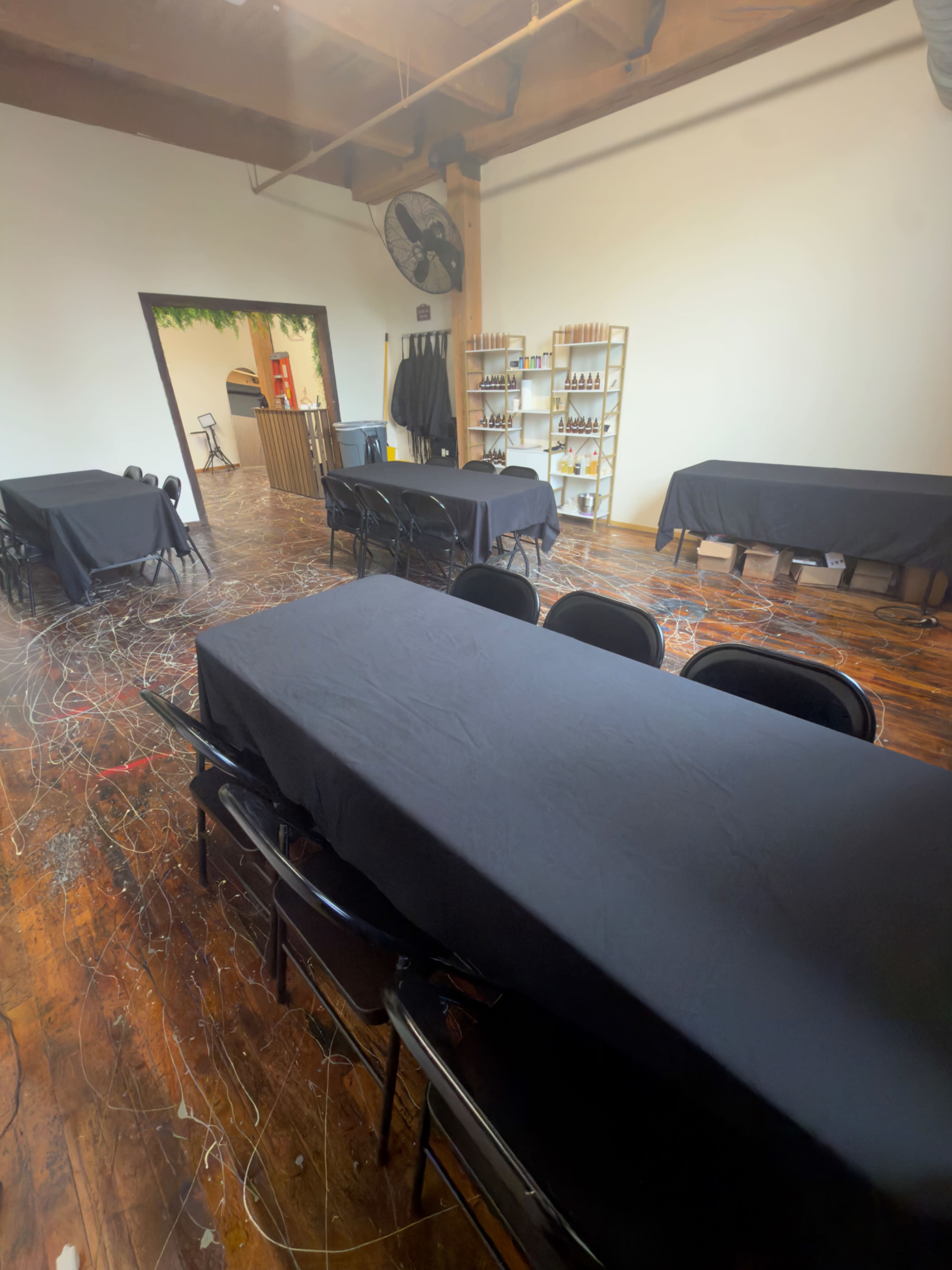 An empty room features several rectangular tables covered with black tablecloths, set against a backdrop of wooden floors and exposed beams.