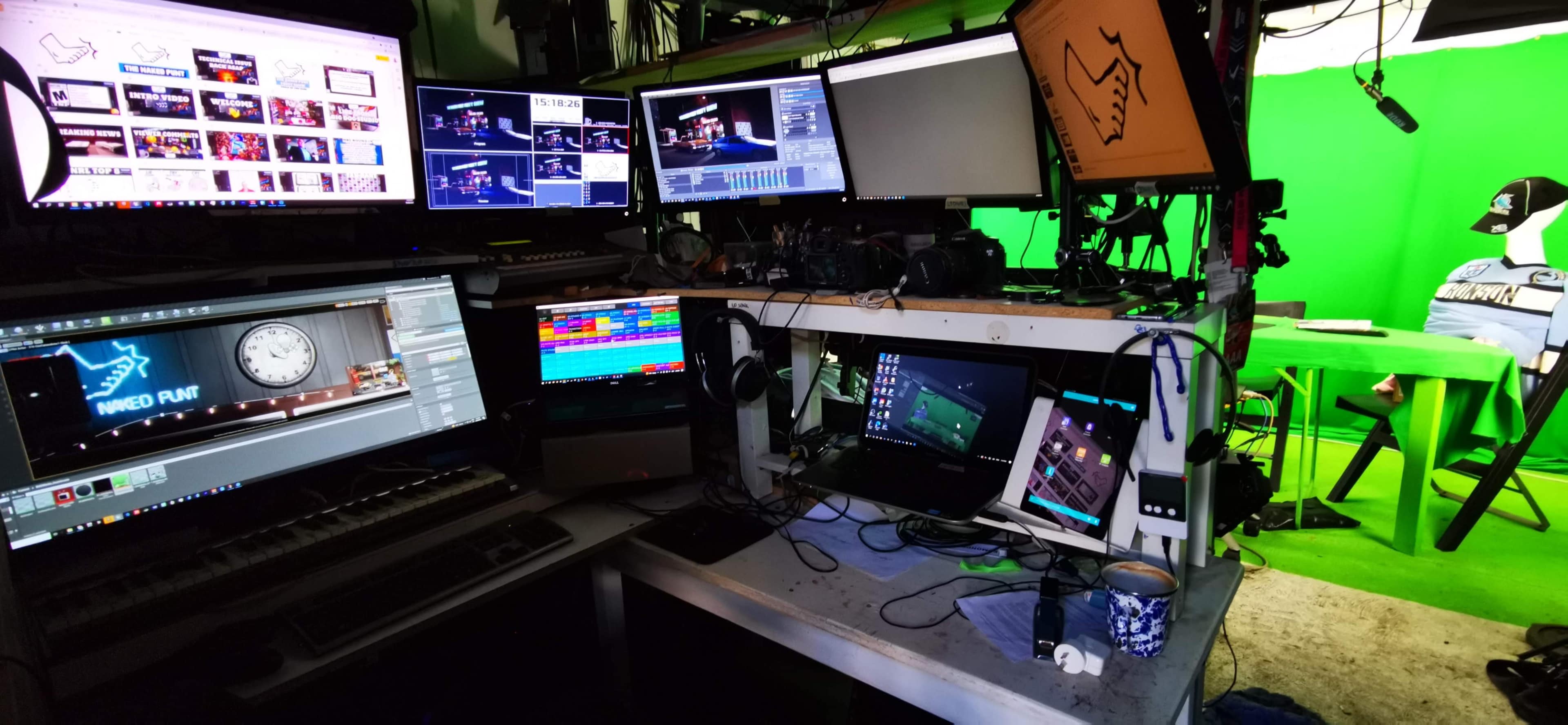 A cluttered multimedia workspace with multiple computer monitors, a green screen backdrop, and various camera equipment.