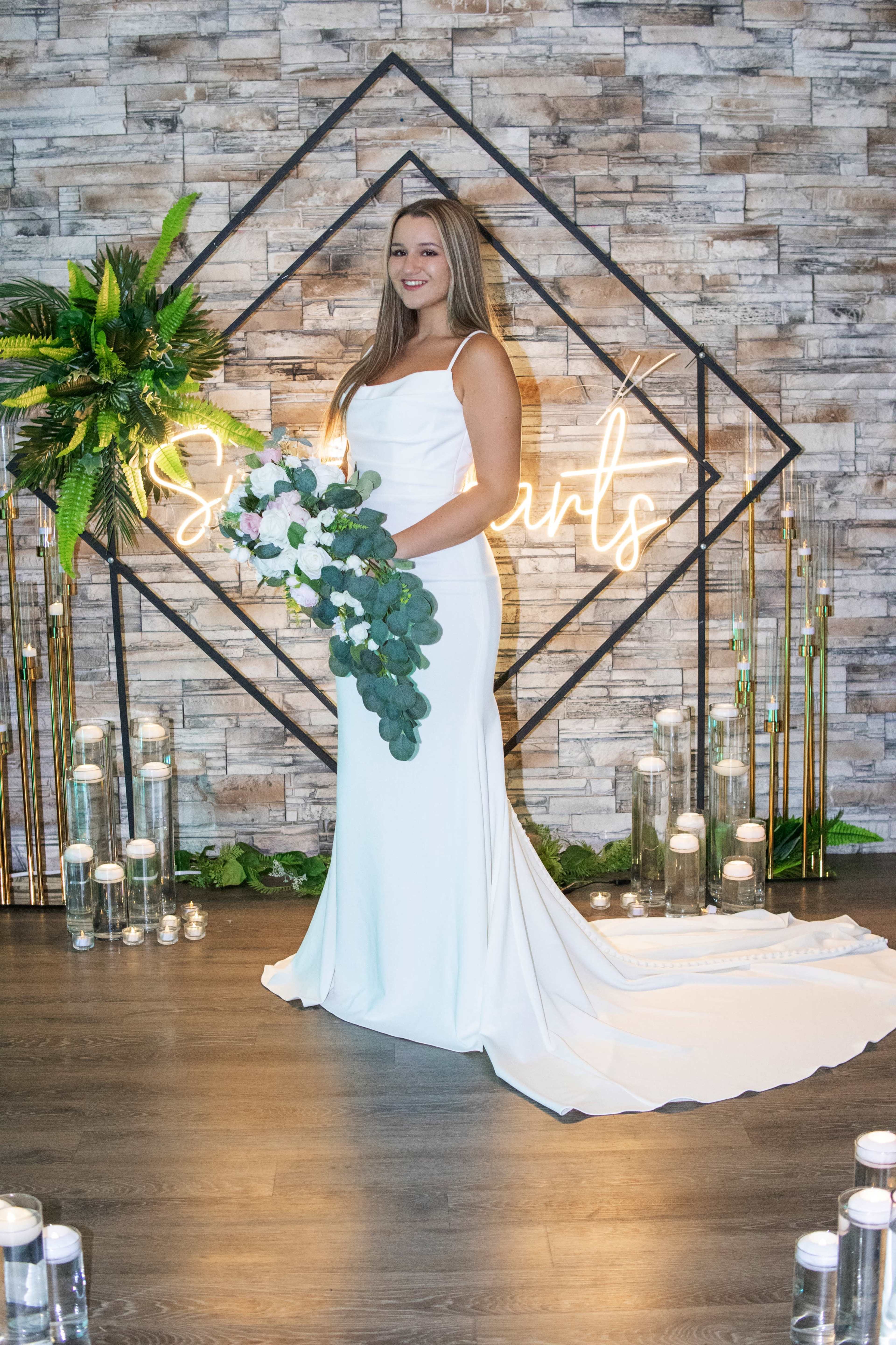 A woman in a white wedding gown stands in front of a decorative backdrop featuring a neon sign and candles.