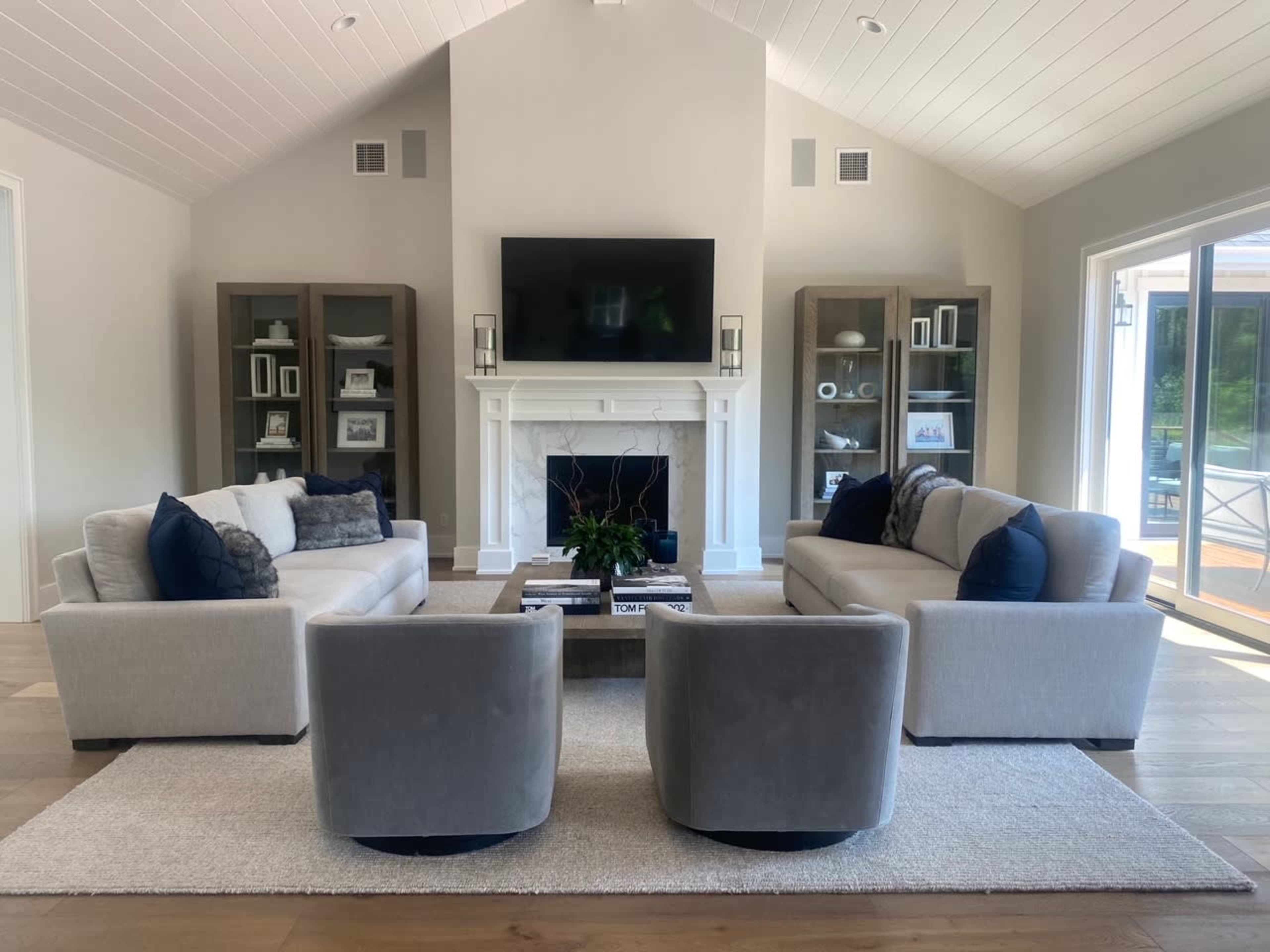 A modern living room featuring two gray sofas and two matching armchairs arranged around a central coffee table, with a television mounted above a white fireplace.