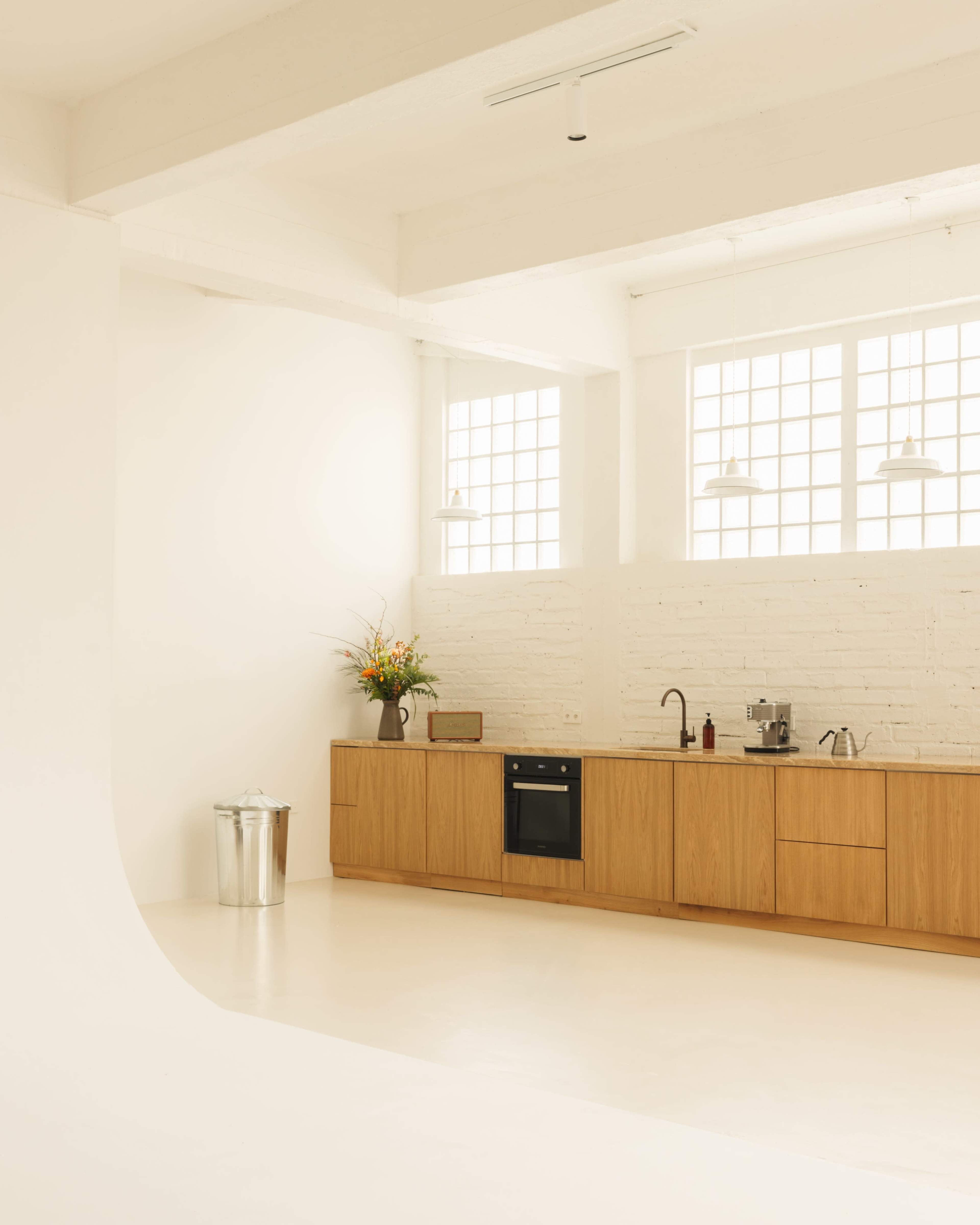 A minimalist kitchen features wooden cabinetry, a sink, a countertop with small appliances, and a window with flower arrangements, set in a bright, white space.