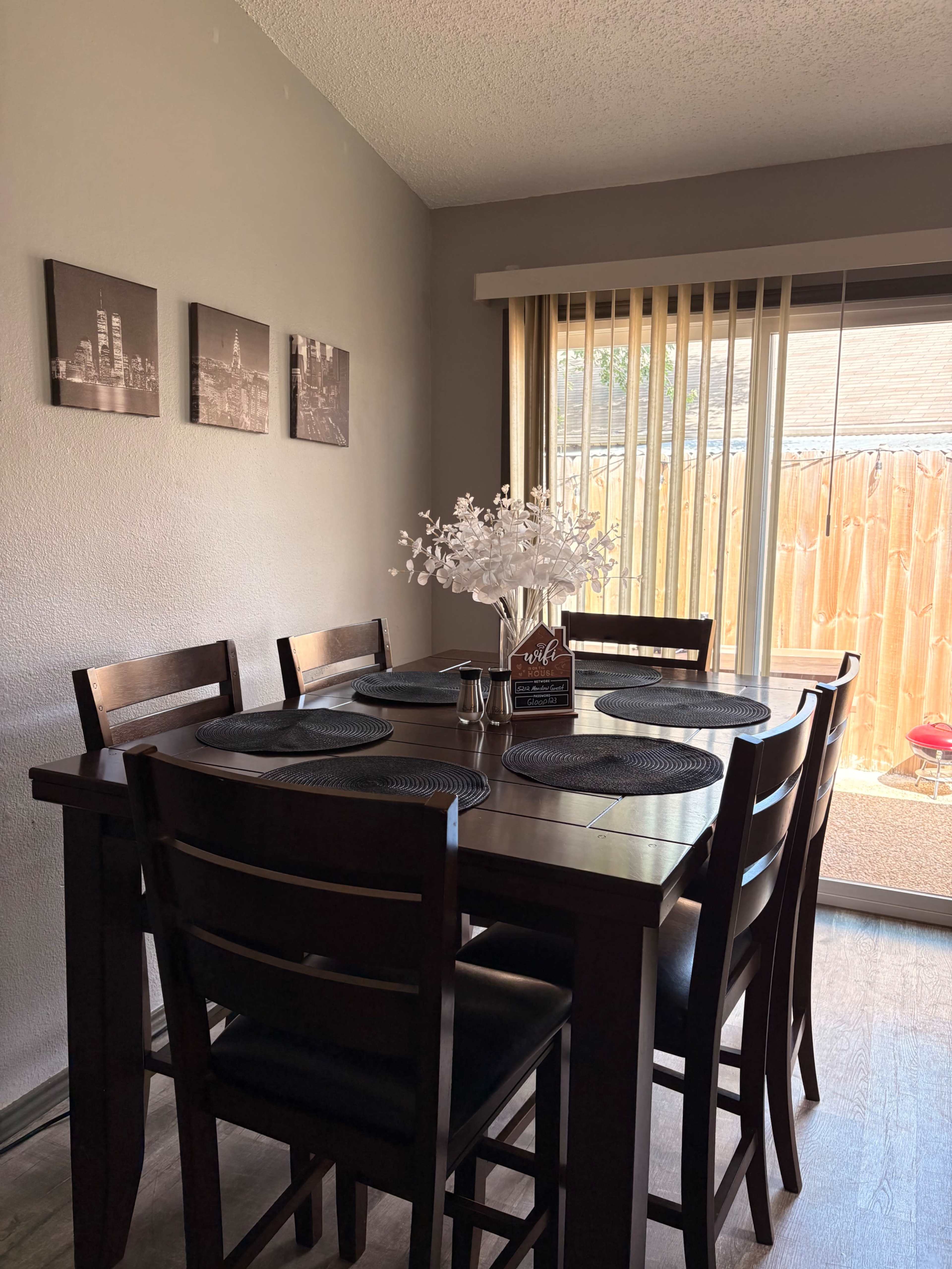 A dining area features a wooden table set for six, with placemats and a vase of flowers, and large sliding glass doors revealing an outdoor space.