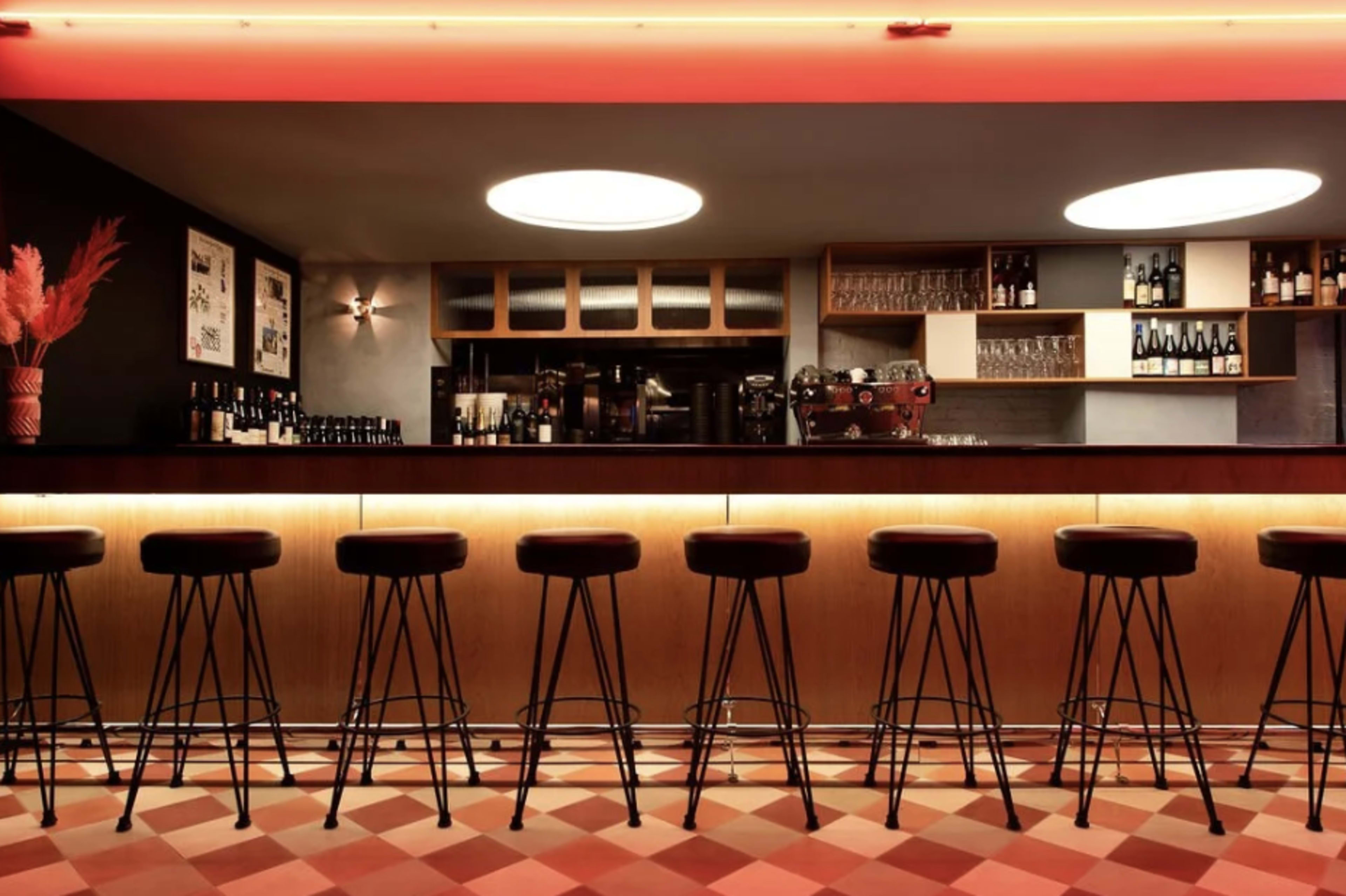 The image shows a modern bar with several black stools lined up in front of a wooden counter, illuminated by red lighting.