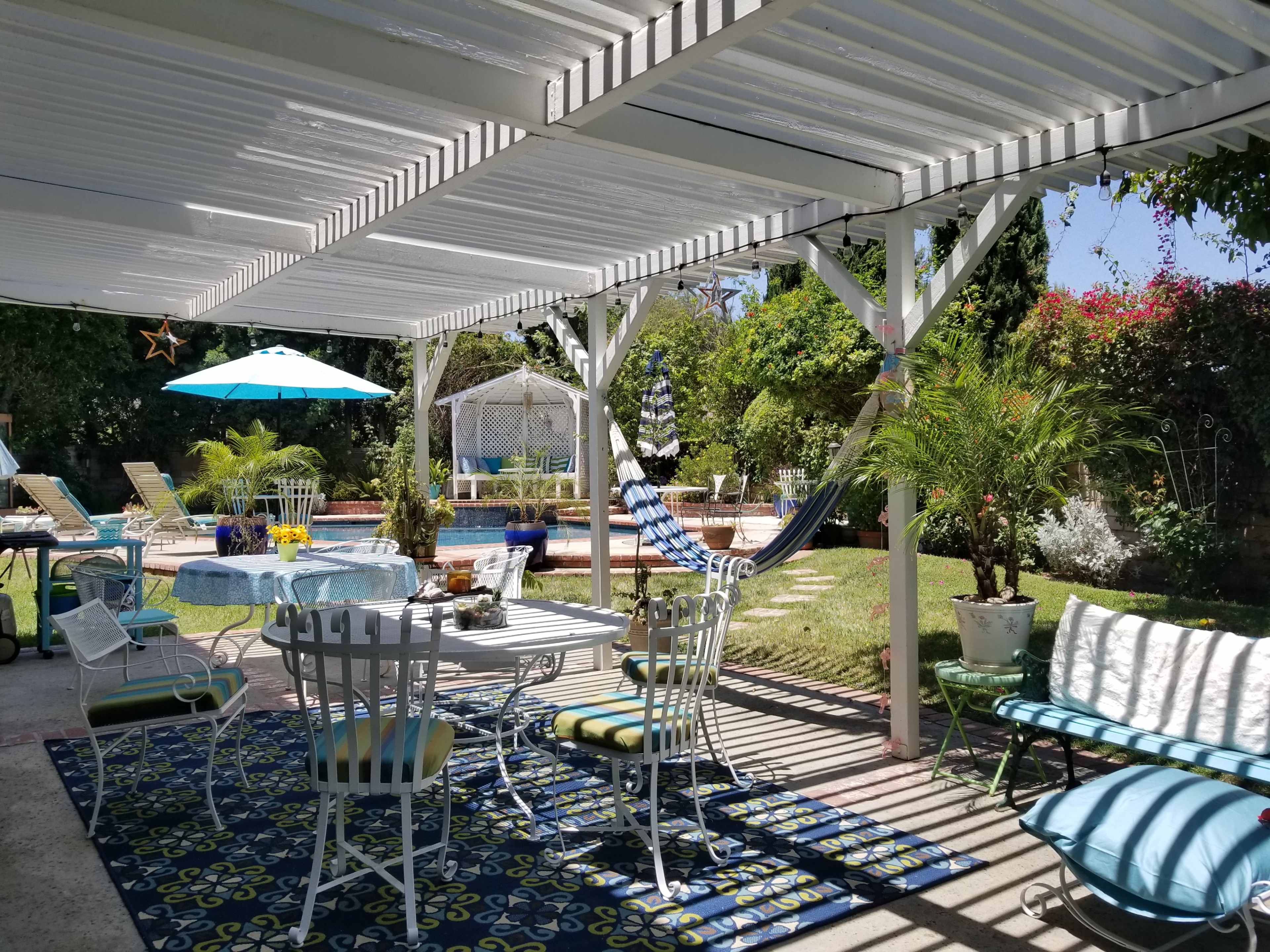 A shaded patio area with a dining table, lounge chairs, and a hammock overlooks a garden and pool.