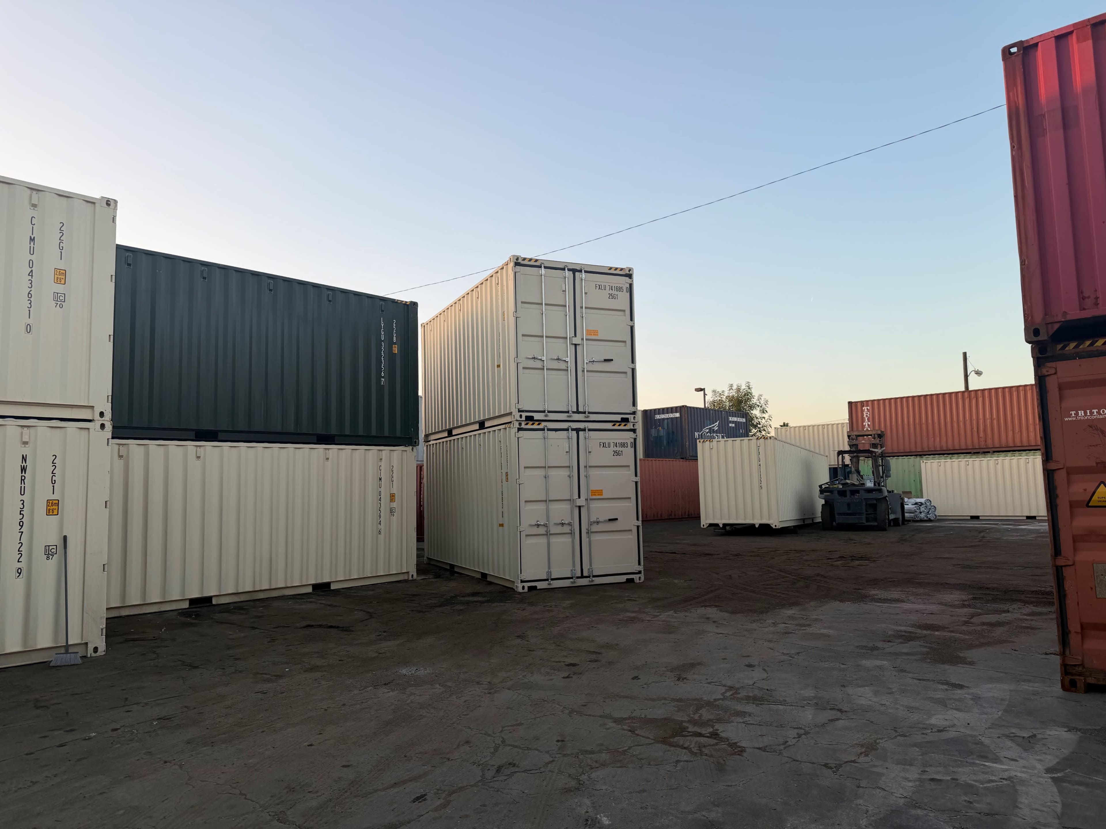 The image shows a storage yard filled with stacked shipping containers of various colors and sizes, illuminated by a clear sky at dusk.