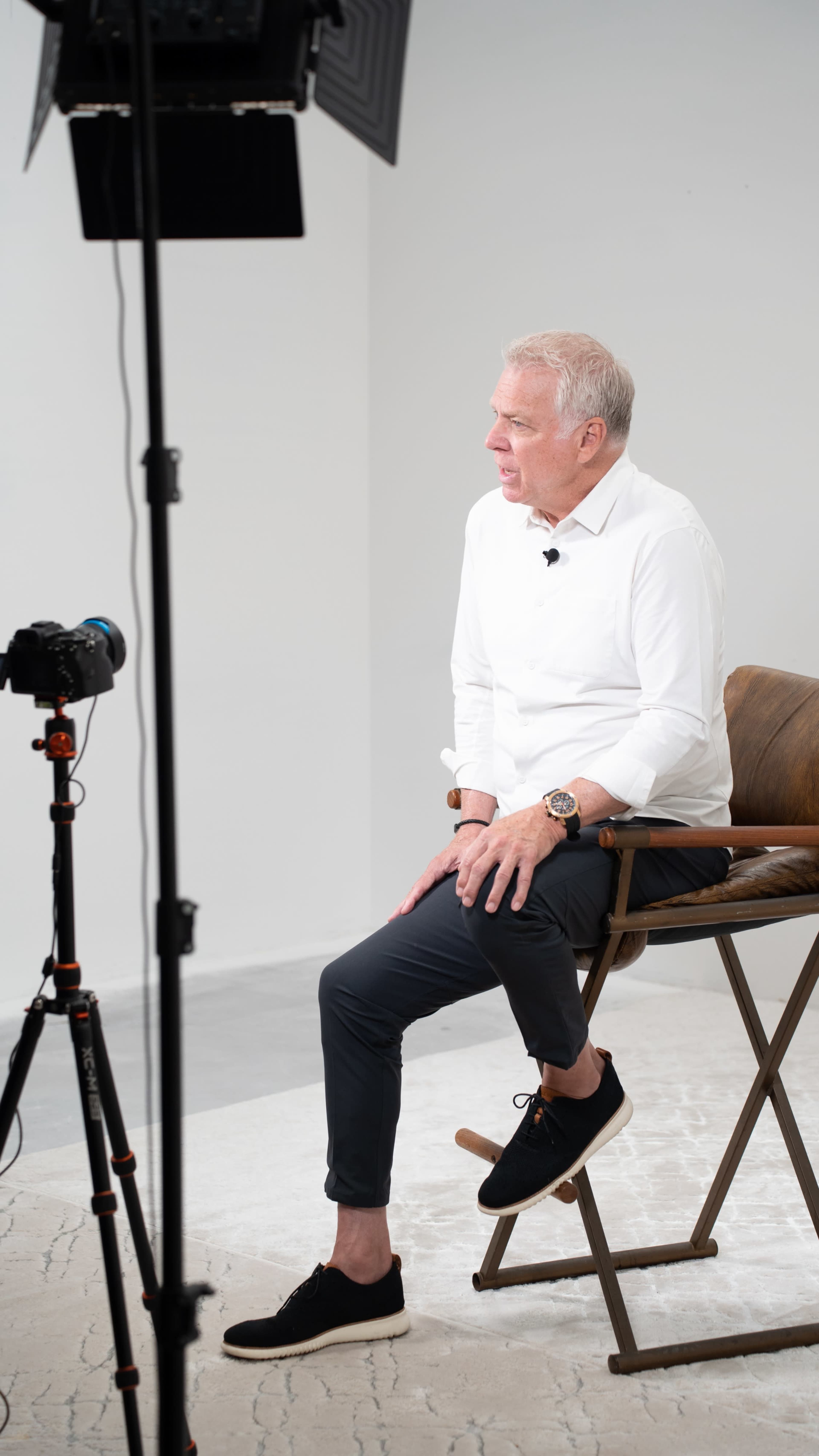 A man in a white shirt sits on a wooden chair in front of a camera on a tripod in a simple, well-lit studio.